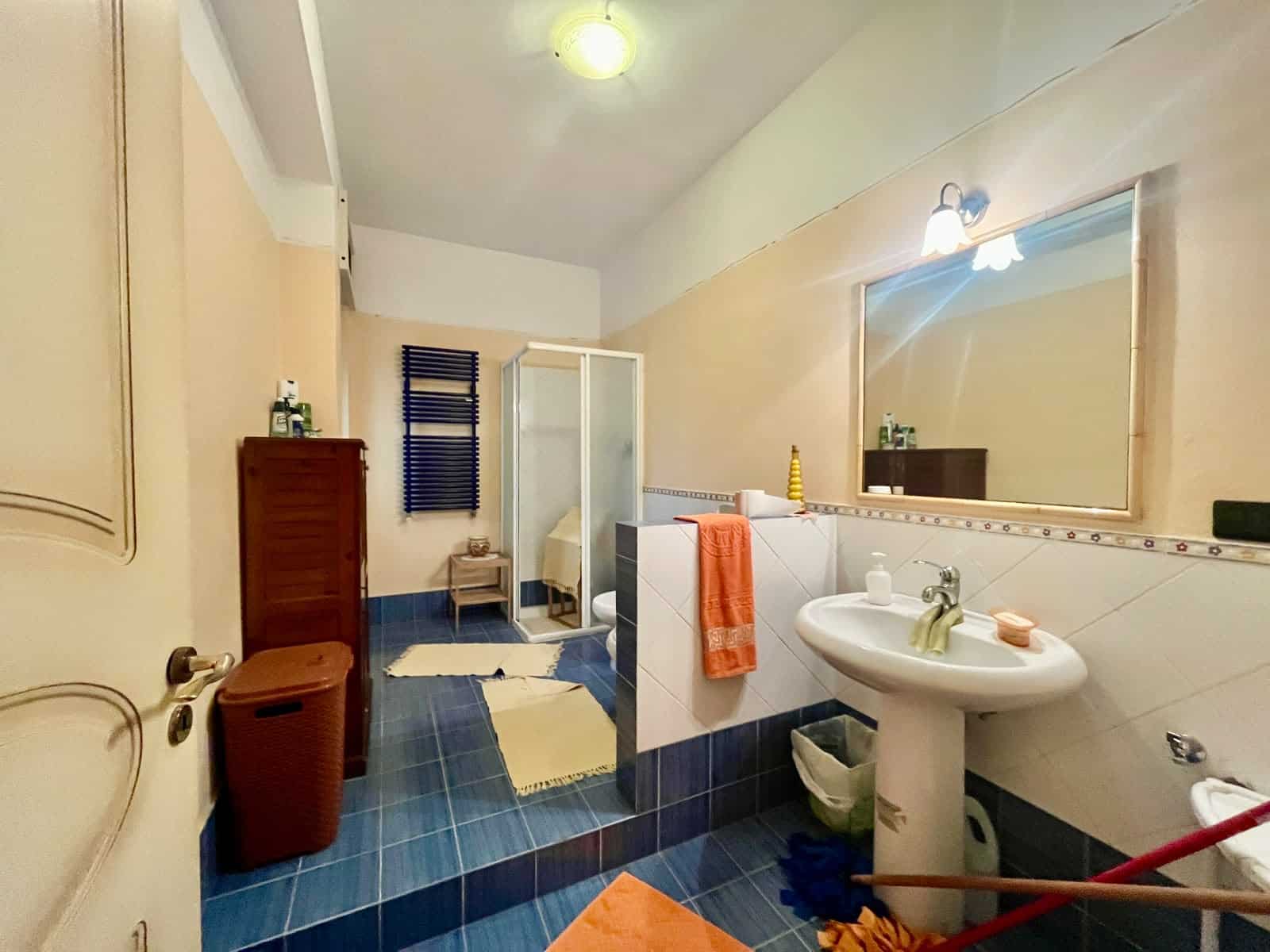 Doccia moderna con pareti in vetro e piastrelle blu, bagno spazioso con lavabo, specchio grande e accessori eleganti, illuminazione soffusa, ideale per comfort e stile.