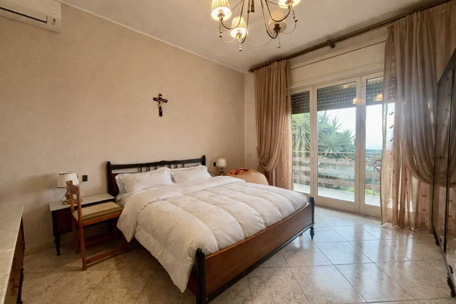 Letto matrimoniale in camera da letto con vista su balcone e paesaggio urbano, arredamento semplice e luminoso, con tende beige e pareti chiare, ideale per soggiorno confortevole e rilassante.