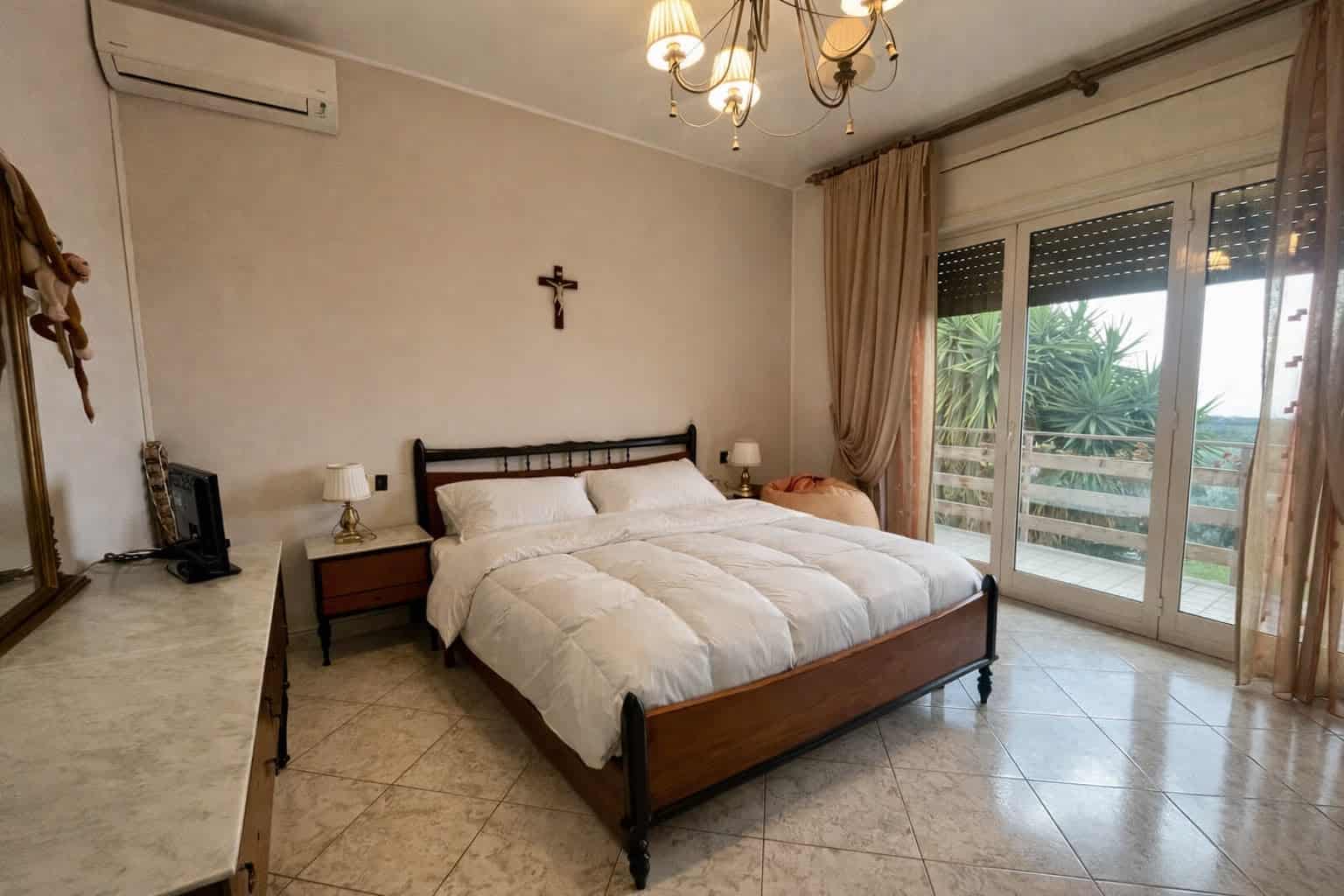Camera da letto spaziosa con letto matrimoniale, armadio in legno, lampade da comodino, e balcone con vista su piante tropicali, in un ambiente luminoso e accogliente.
