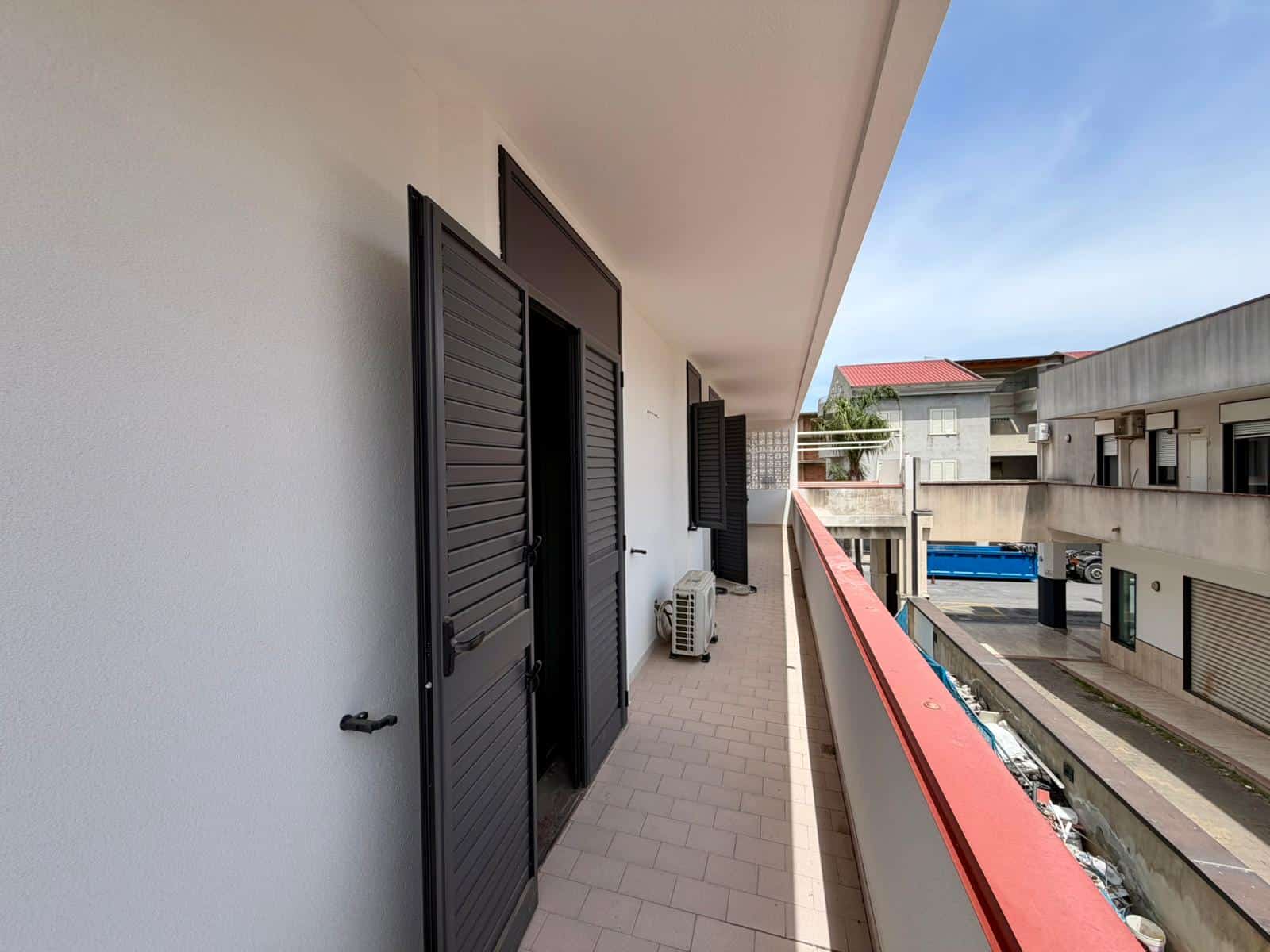 Balcone di un appartamento con porte a persiana nere, vista su strada urbana con edifici moderni, cielo sereno e ambiente residenziale. Ideale per immobili in affitto o vendita a NextCasa.