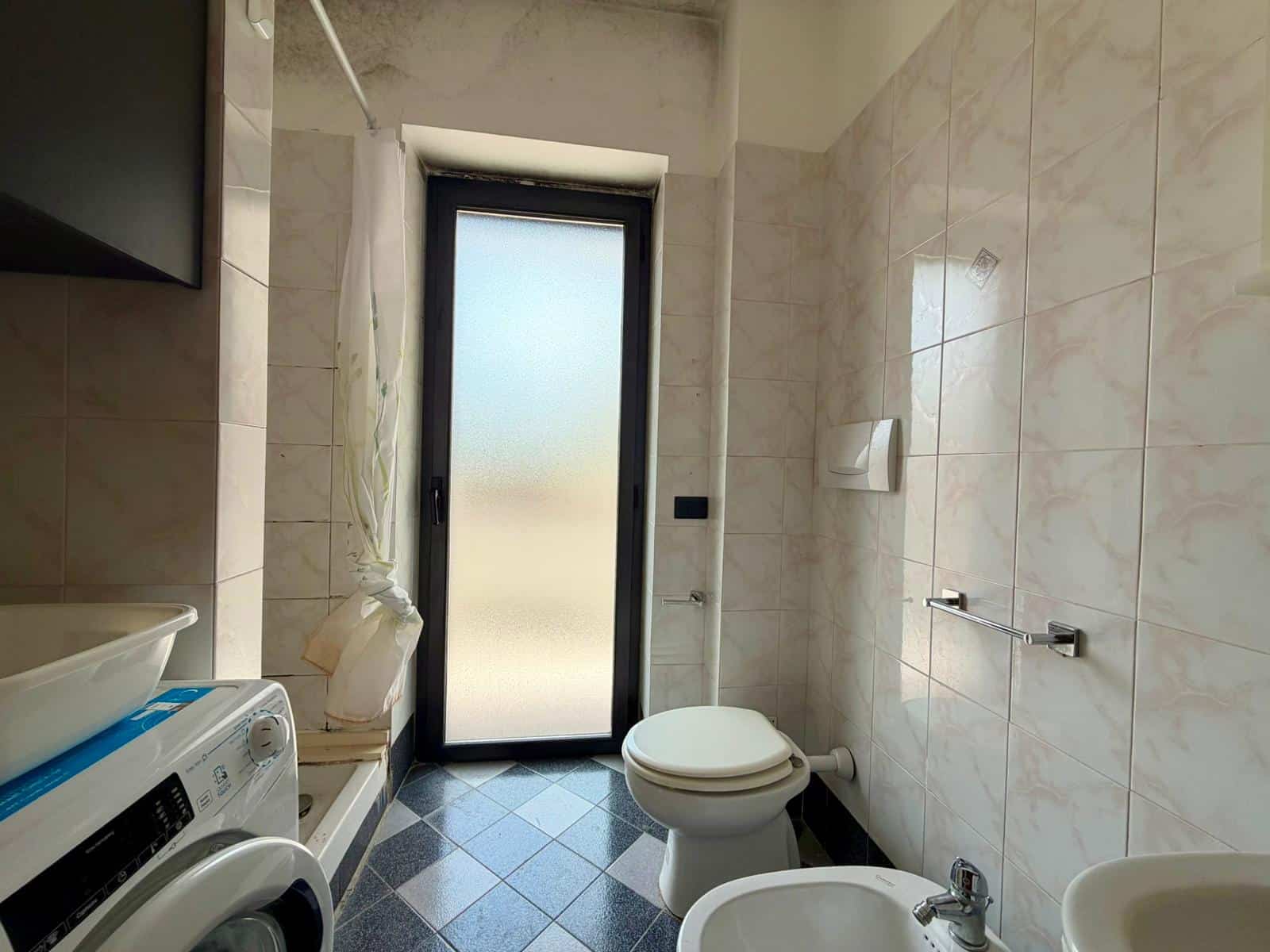 Lavabo e servizi igienici moderni in un bagno luminoso con pareti in piastrelle beige e porta finestra con vetro opaco, ideale per ambienti di comfort e funzionalità.