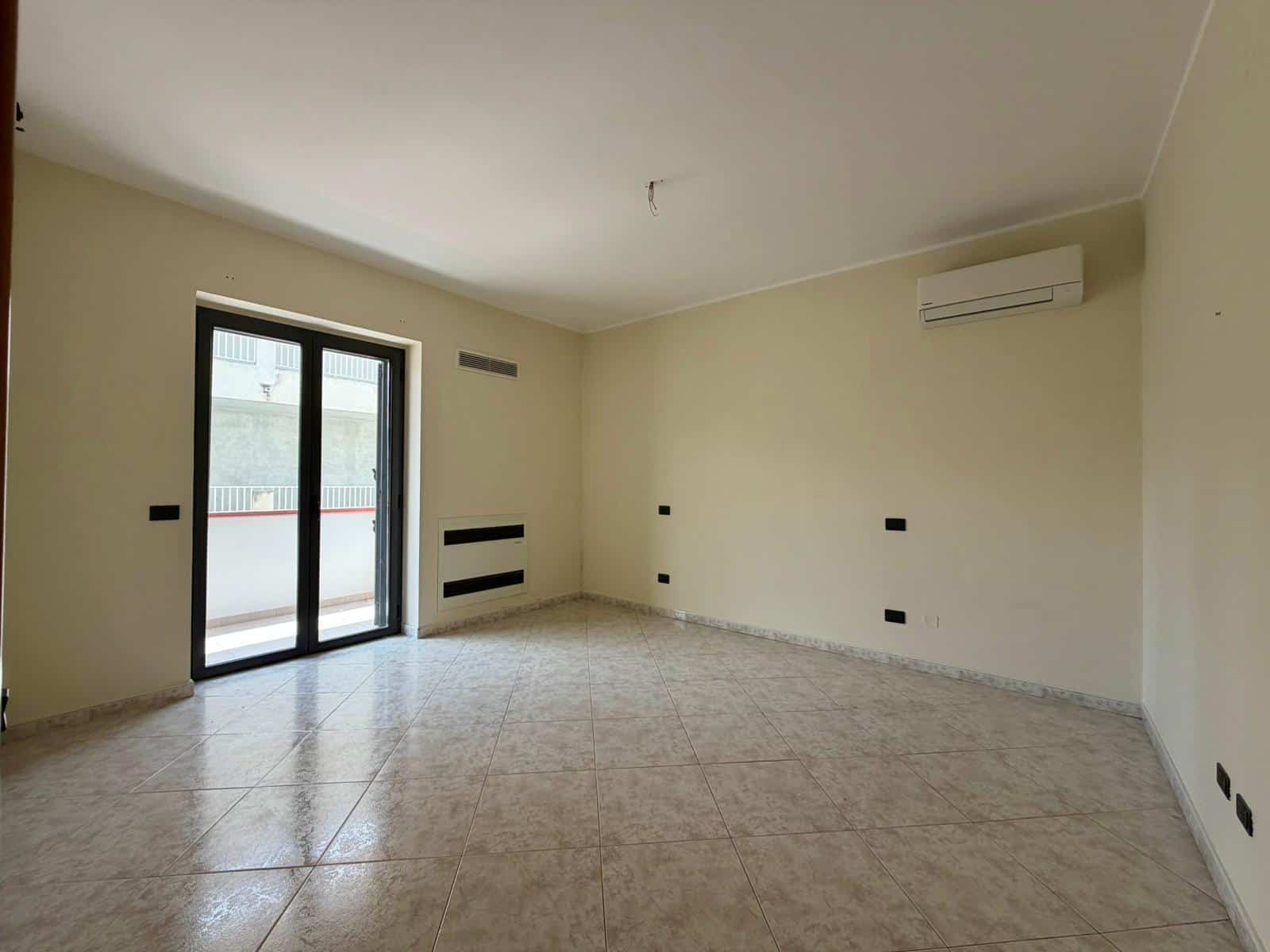 Ampio soggiorno luminoso con pareti neutre, grande finestra e accesso al balcone, ideale per living e relax, dotato di aria condizionata e finiture moderne.