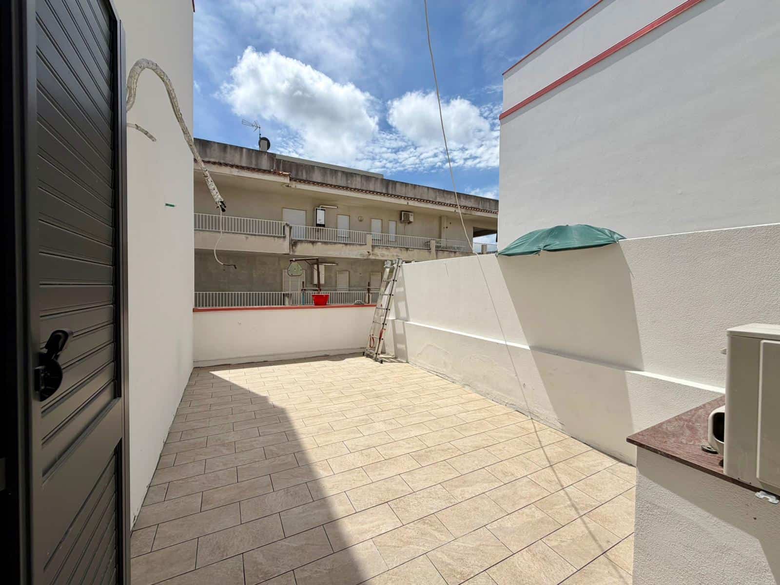 Ampio terrazzo esterno con pavimentazione in piastrelle, pareti bianche e vista sul cielo azzurro con nuvole, ideale per relax e attività all'aperto, in un contesto residenziale moderno.