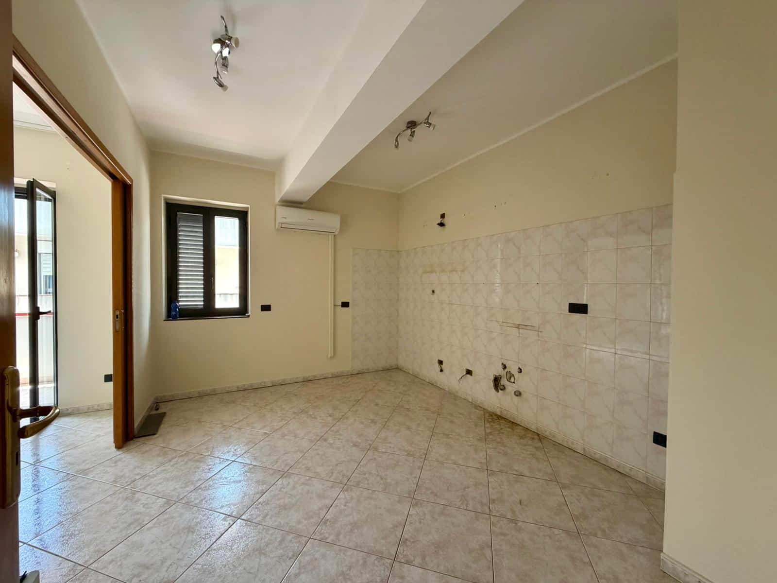 Cucina vuota in fase di ristrutturazione con pareti beige, piastrelle chiare e finestre con persiane, pronta per essere arredato.