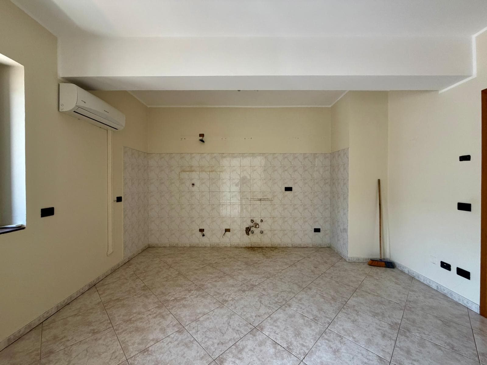 Ampia stanza vuota in fase di ristrutturazione, con pareti beige, piastrelle chiare e aria condizionata, pronta per essere personalizzata come soggiorno o ufficio.