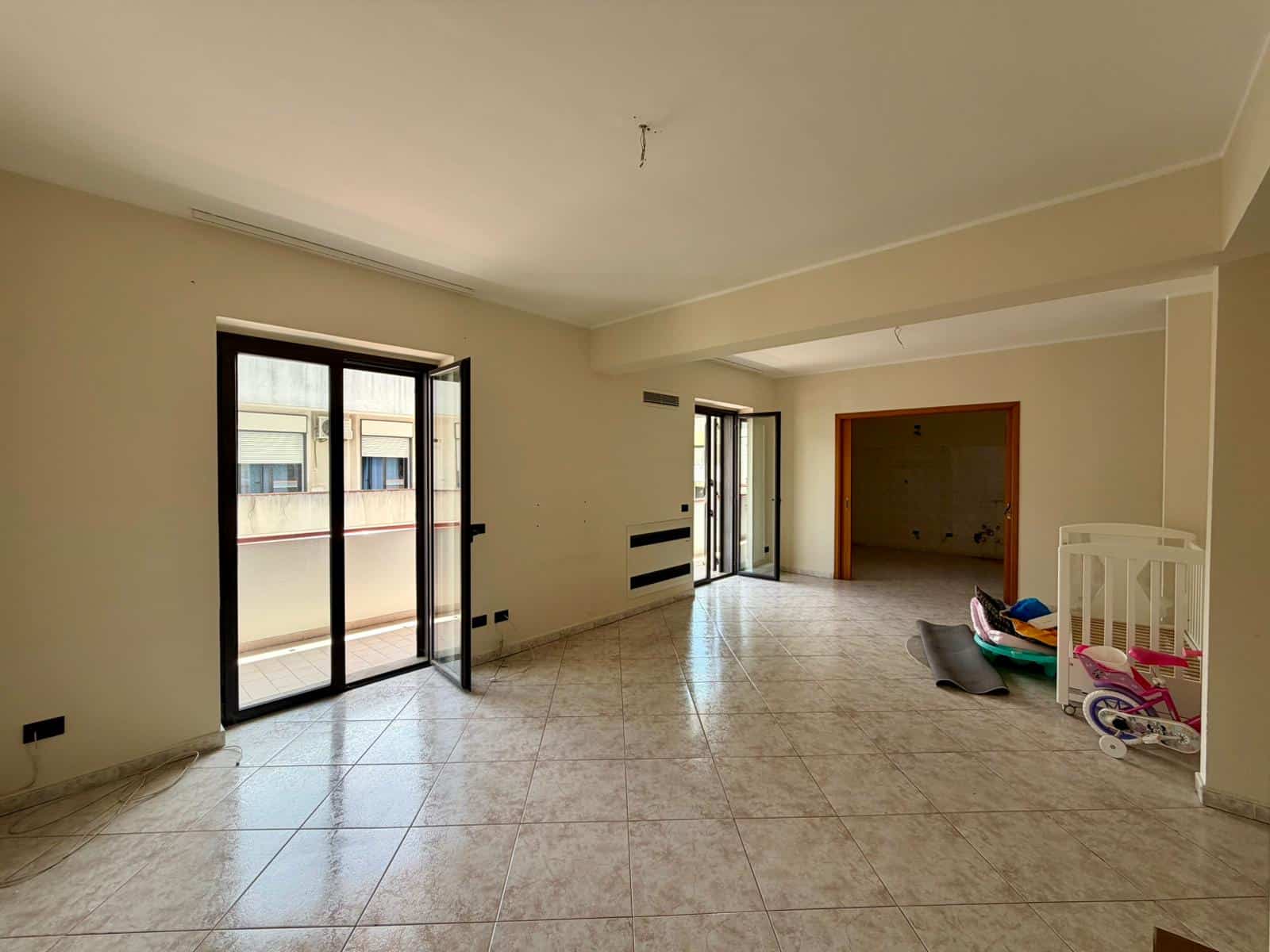 Interno di appartamento vuoto con grandi finestre e accesso al balcone, pavimento in piastrelle chiare, pronto per arredare, in zona residenziale.