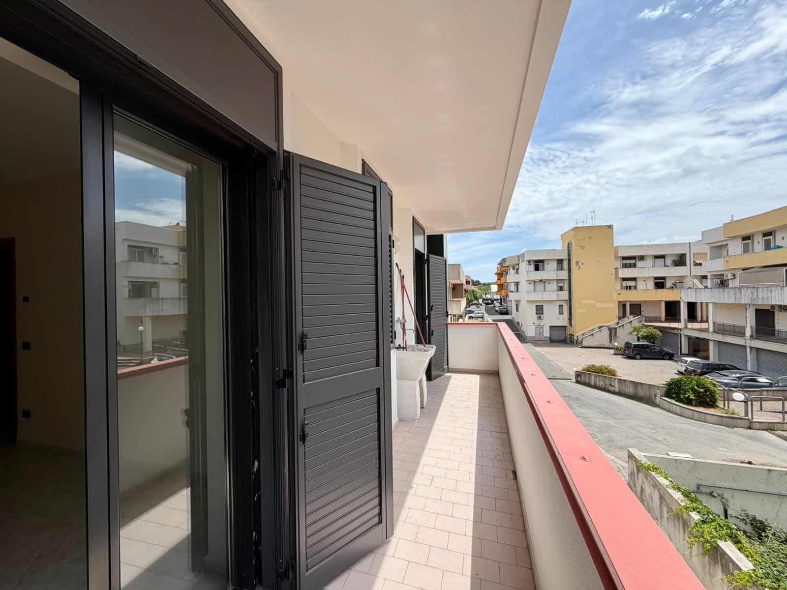 Balcone di un appartamento urbano con vista su edifici residenziali e strada, con parapetto rosso e pavimentazione in piastrelle beige, in un contesto di città moderna e vivace.