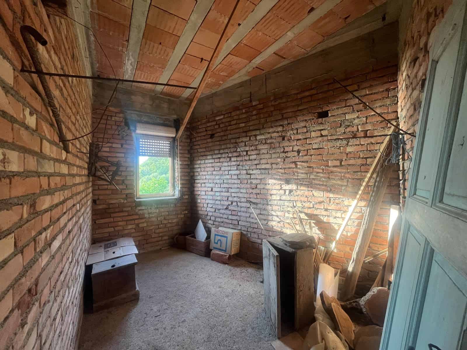 Interno di una stanza in costruzione con pareti in mattoni e soffitto in legno, finestra con vista su natura.