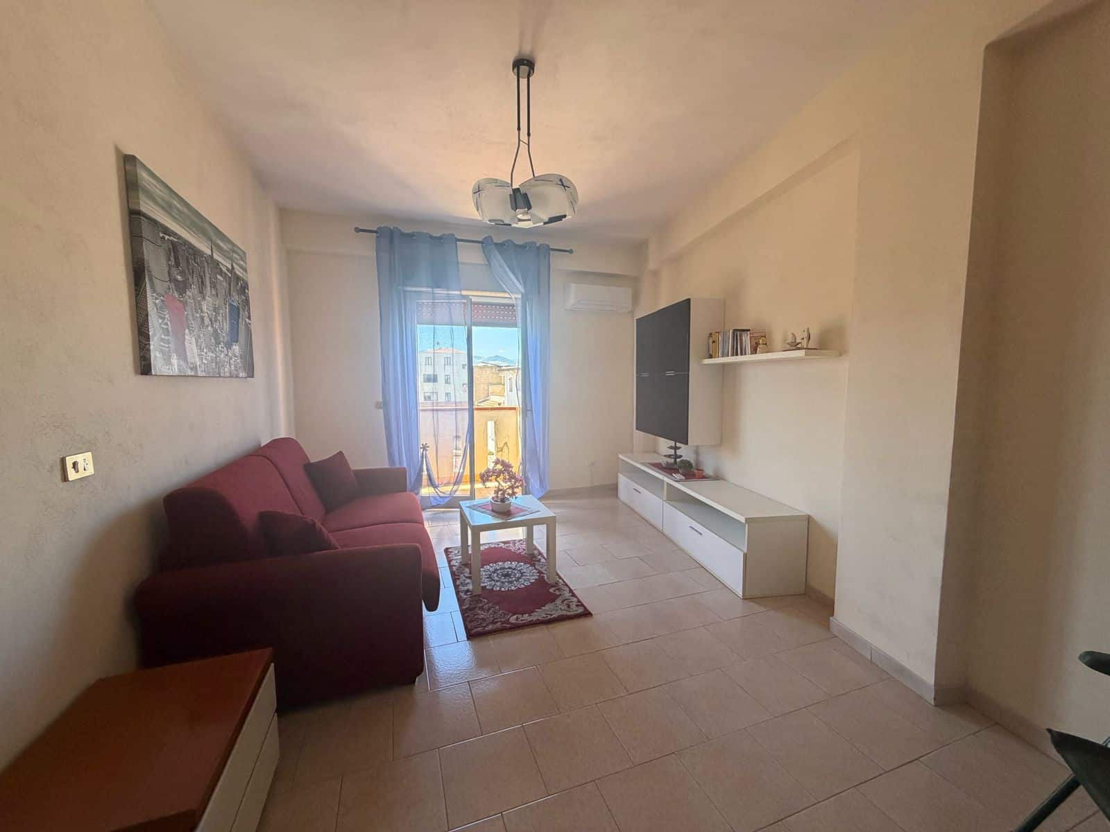 Soggiorno luminoso con divano rosso, TV e balcone con vista sulla città, arredato in stile moderno.