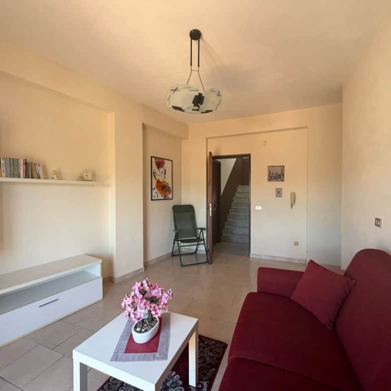 Interno di un appartamento con divano rosso, tavolino con fiori e pareti decorate, ambiente luminoso e accogliente.