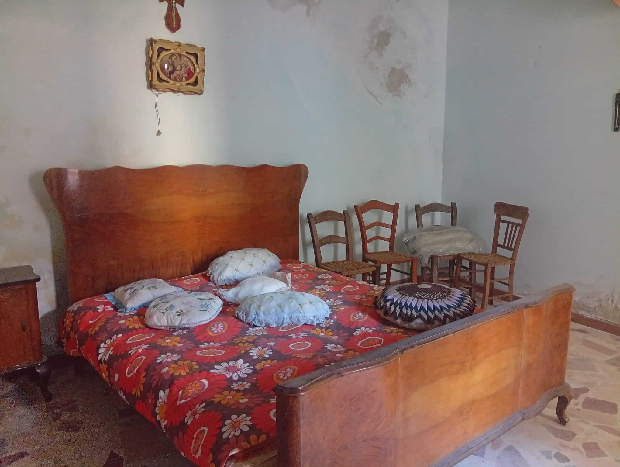 Camera da letto con letto in legno, coperta floreale e quattro cuscini, pareti con segni di umidità e decorazioni semplici.