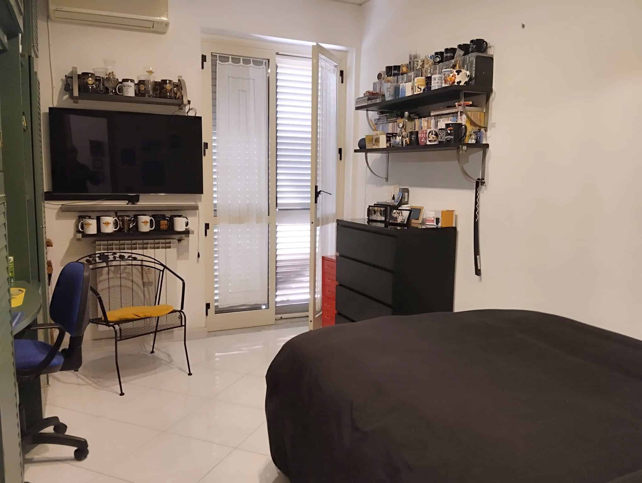 Camera da letto con TV, scaffali con oggetti e porta finestra con persiane in legno.