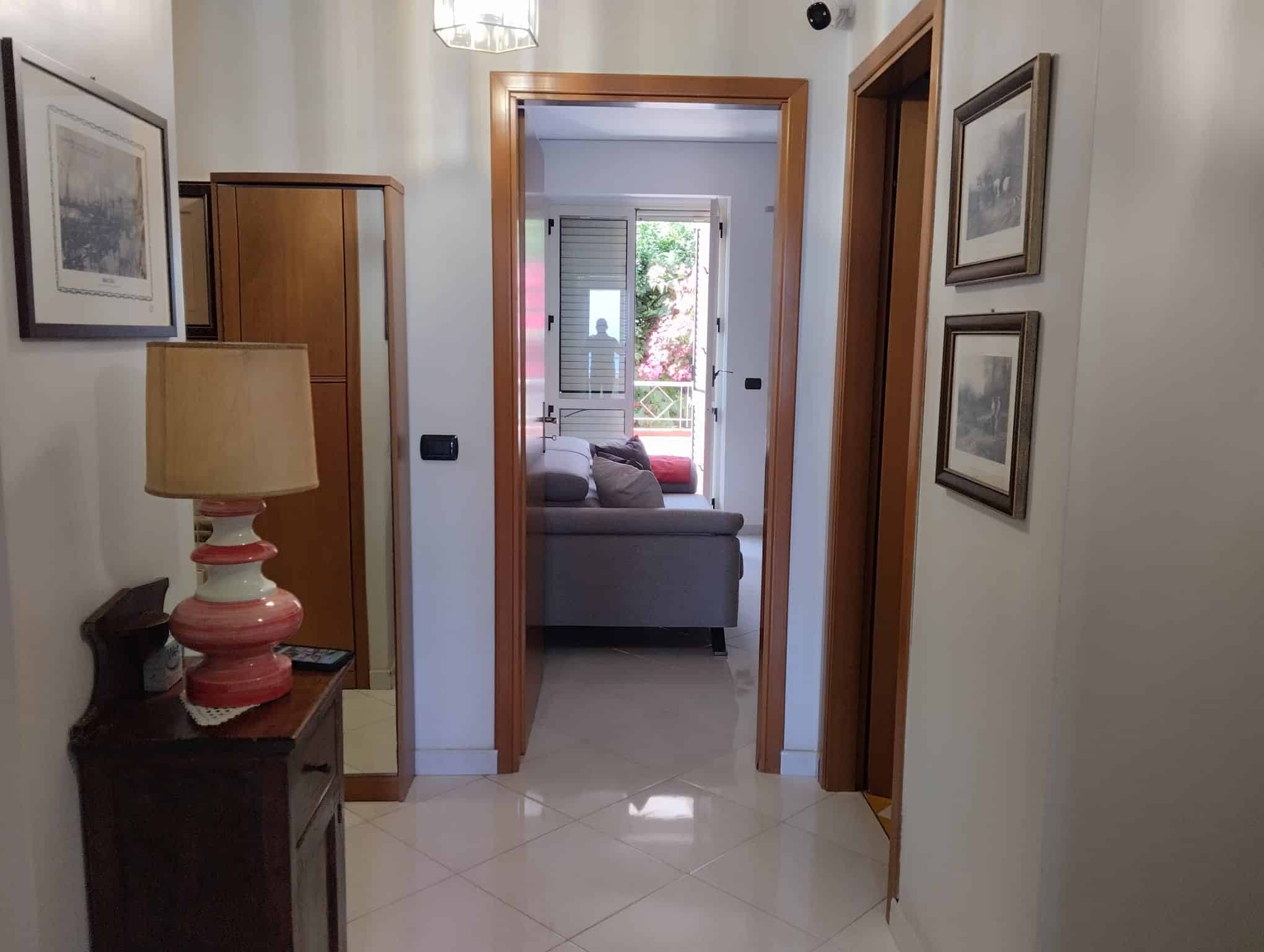Ingresso di un appartamento con pareti bianche, quadri alle pareti e mobili in legno, vista sulla zona giorno con divano e finestra con vista sul balcone.
