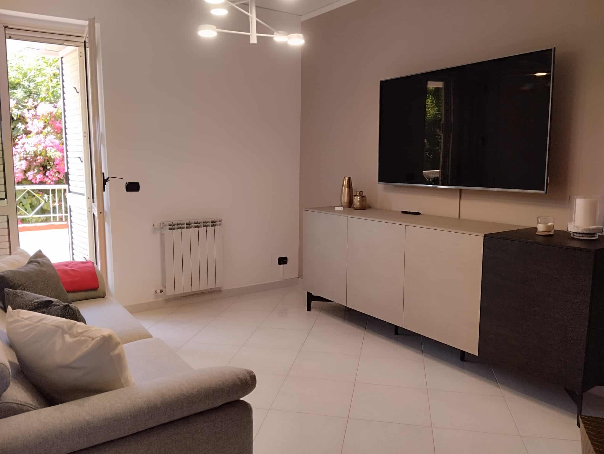 Interno di un soggiorno moderno con divano, TV a schermo piatto e accessori decorativi, luce naturale e balcone con vista.
