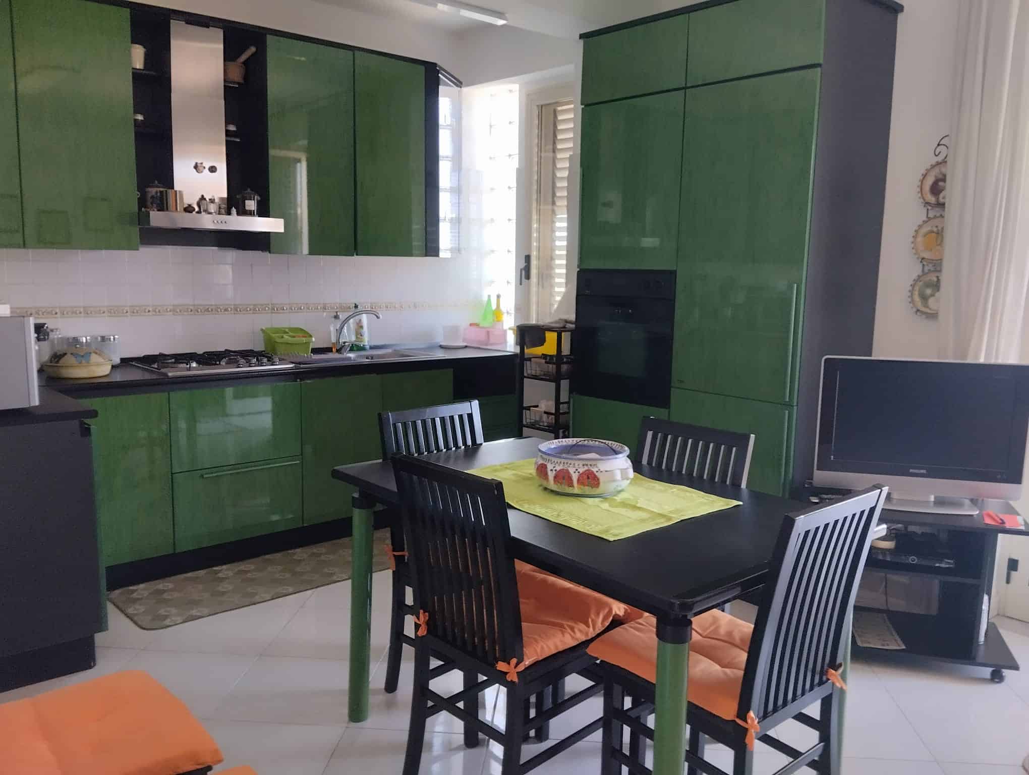 Cucina moderna con mobili verdi, tavolo nero e sedie con cuscini arancioni, ambiente luminoso e accogliente.