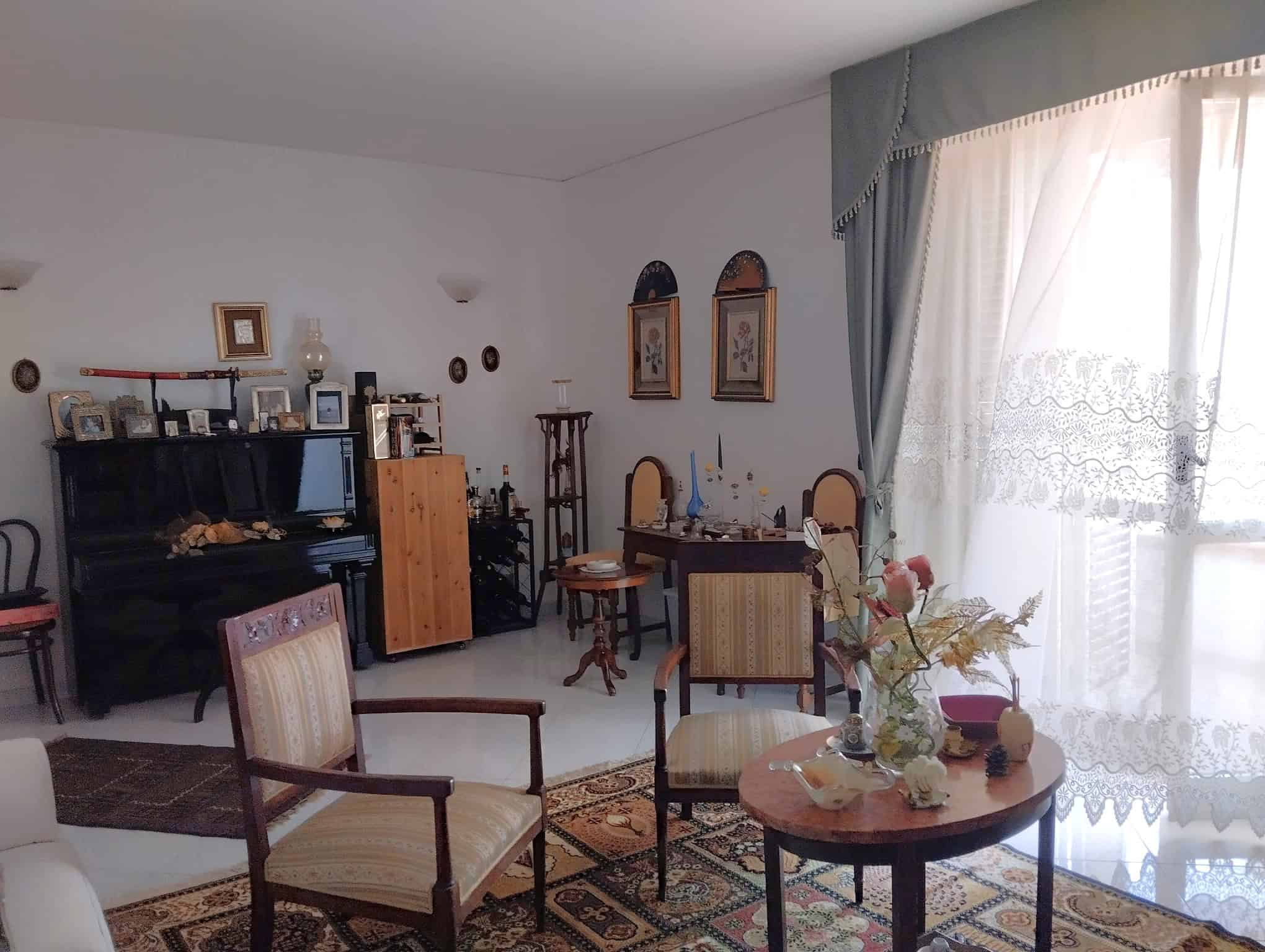 Interno di un soggiorno con mobili d'epoca, tappeti e decorazioni classiche, illuminato da luce naturale attraverso una grande finestra con tende.