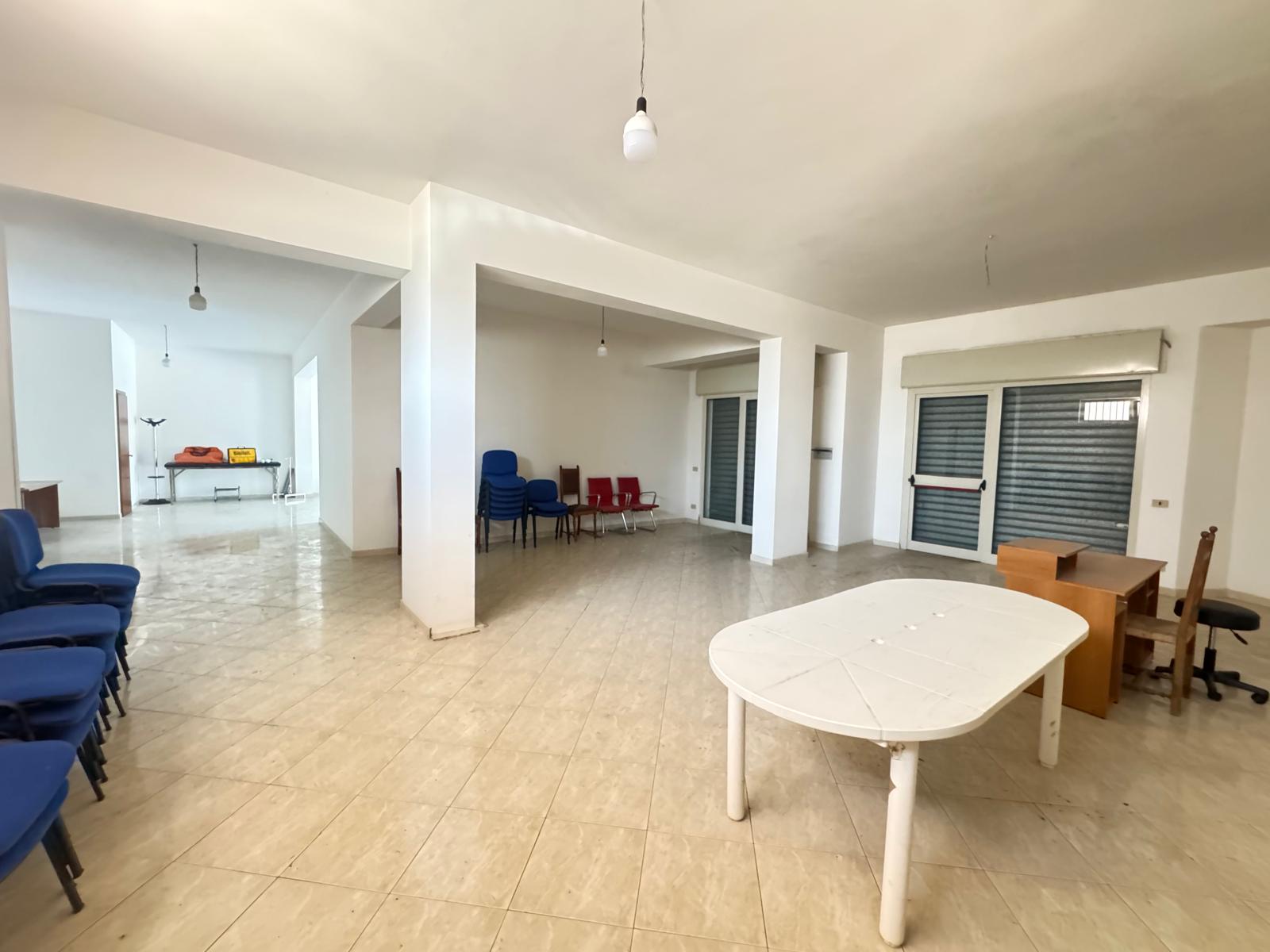 Spazioso ufficio o sala riunioni con grandi finestre, tavolo bianco e sedie blu e rosse, ambiente luminoso e arioso.