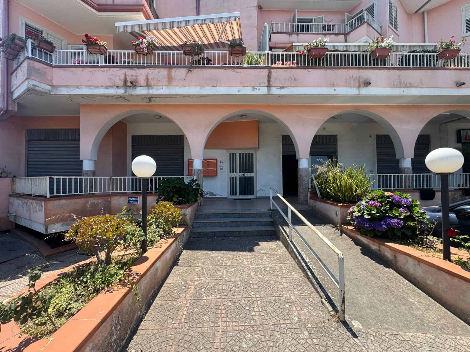 Ingresso di un edificio residenziale con rampe e giardini ben curati, in stile mediterraneo, con balconi e fiori colorati, ideale per immobili in vendita o affitto a NextCasa.