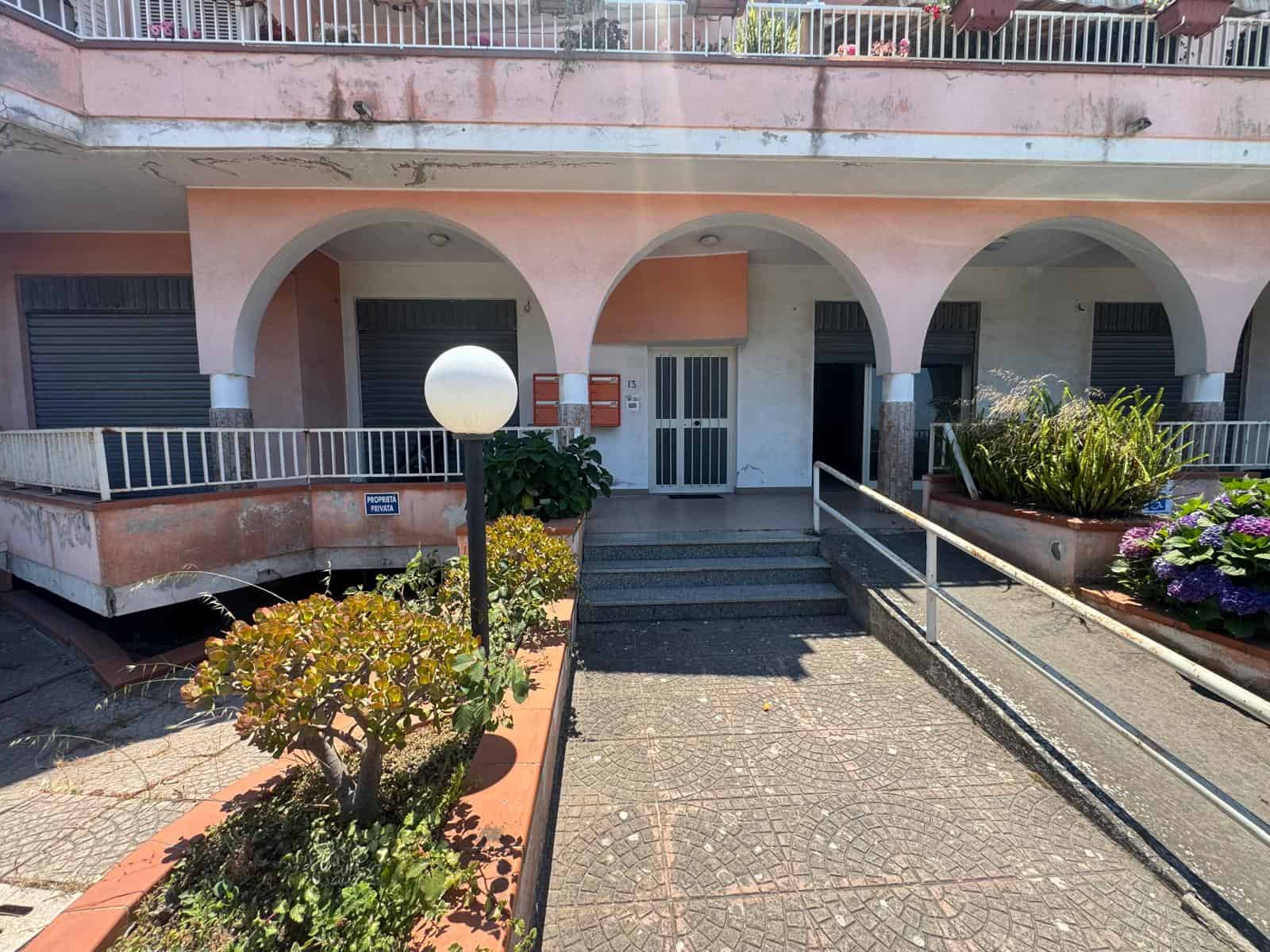 Ingresso di un edificio residenziale con rampe e archi, circondato da piante e fiori, in stile mediterraneo.