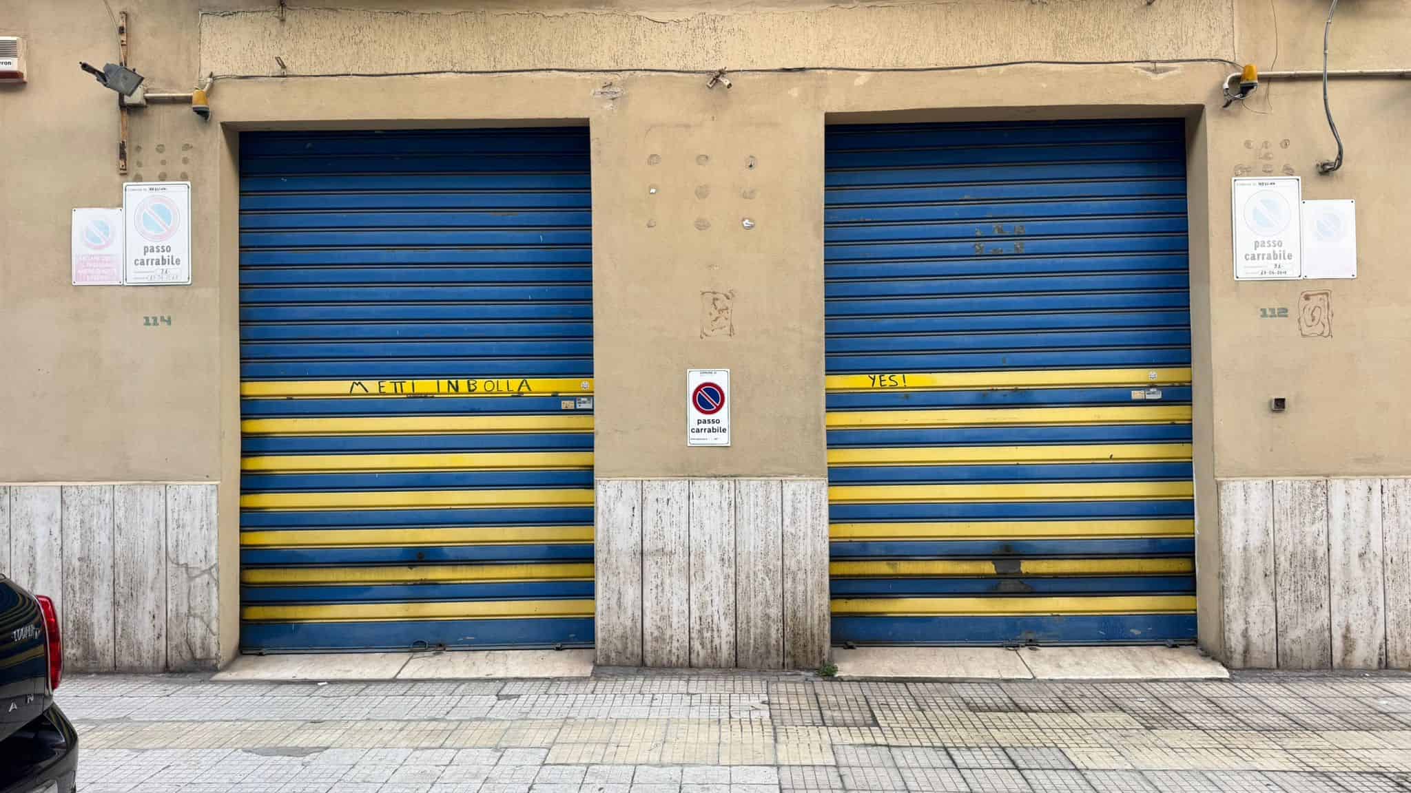 Due portoni industriali con sbarre gialle e blu, situati in un edificio urbano, ideali per magazzini o attività commerciali, con un design funzionale e resistente.