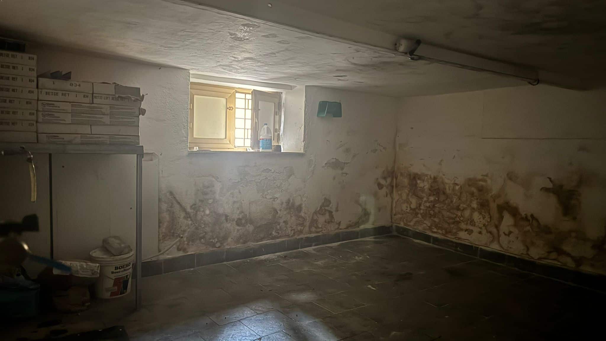 Magazzino con pareti umide e muffa, illuminato da una piccola finestra, in condizioni di degrado e necessità di ristrutturazione.