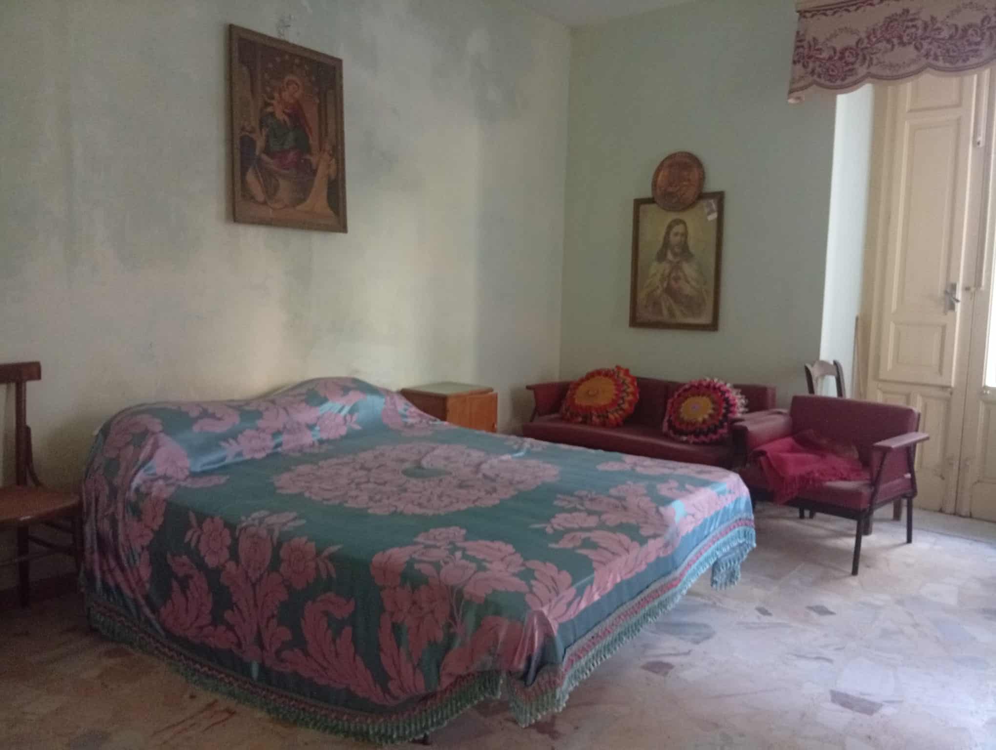 Interno di una camera da letto con letto matrimoniale, divano, sedie e decorazioni religiose sulle pareti in una casa in vendita a Ficarra, Messina.