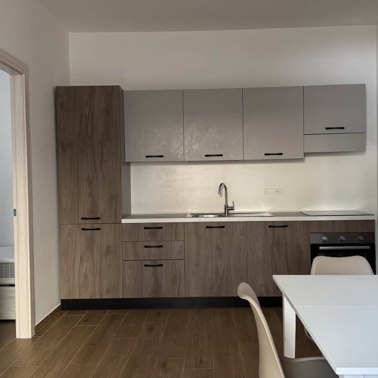 Cucina moderna con mobili in legno chiaro, pareti bianche e piano di lavoro in laminato, dotata di lavello e armadietti superiori e inferiori, ambiente luminoso e funzionale.