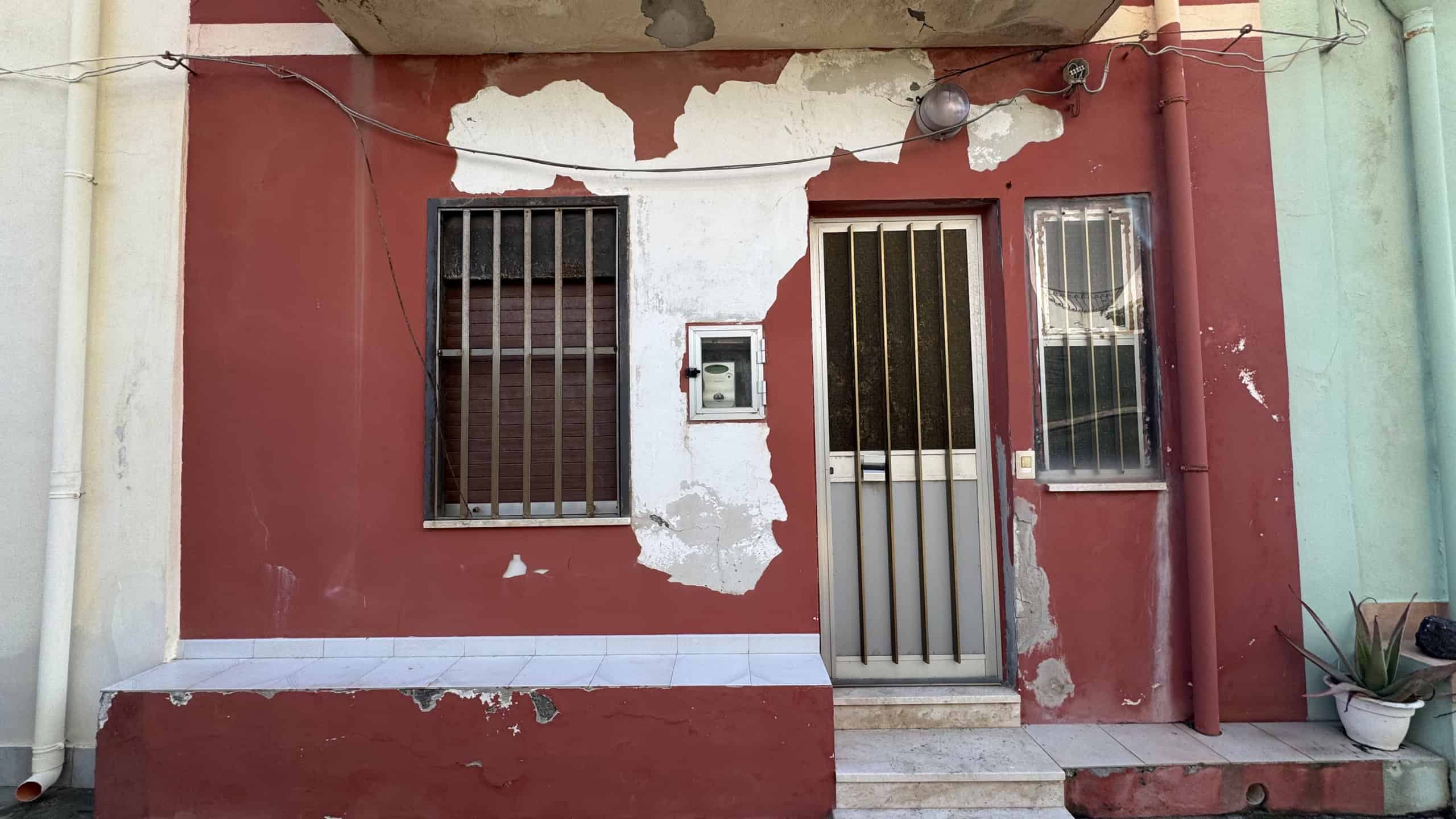 Scorcio di facciata di edificio residenziale con pareti in pietra e infissi in metallo, mostra segni di deterioramento e pittura scrostata, tipico di un'area urbana storica in Italia.