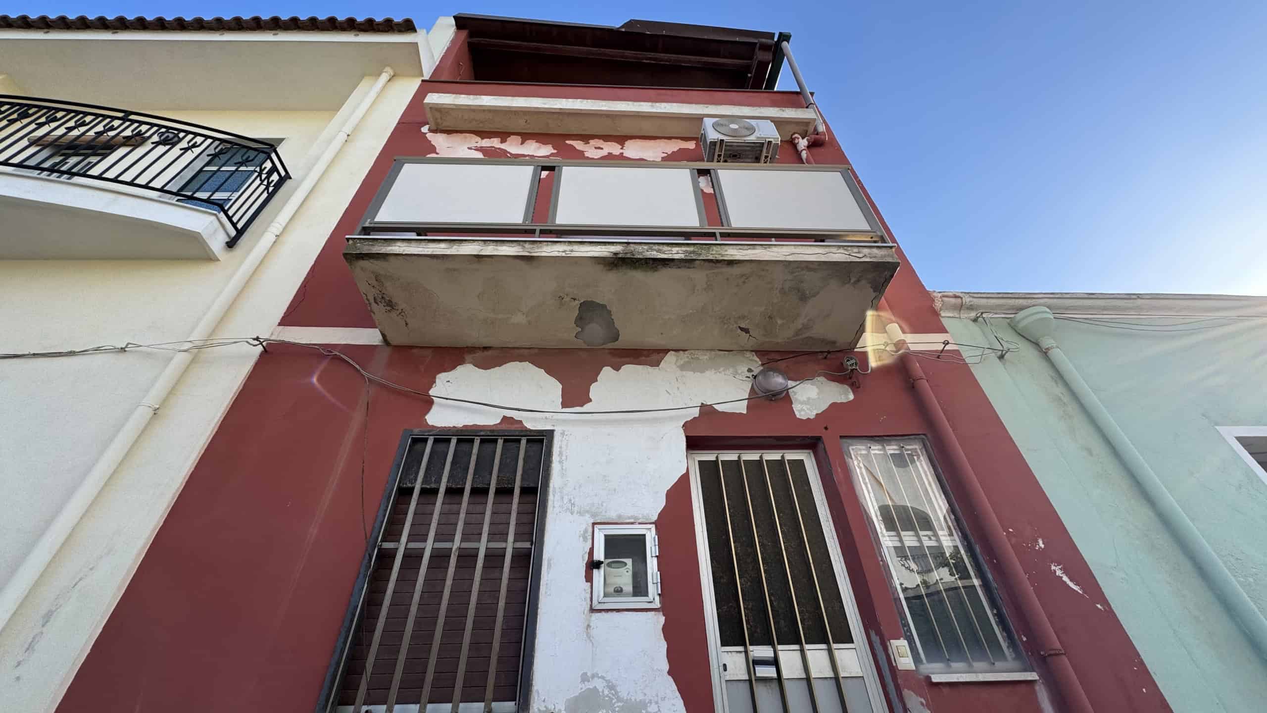 Ampio edificio residenziale con balcone e facciata in condizioni di deterioramento, situato in un contesto urbano con edifici adiacenti, sotto un cielo sereno.