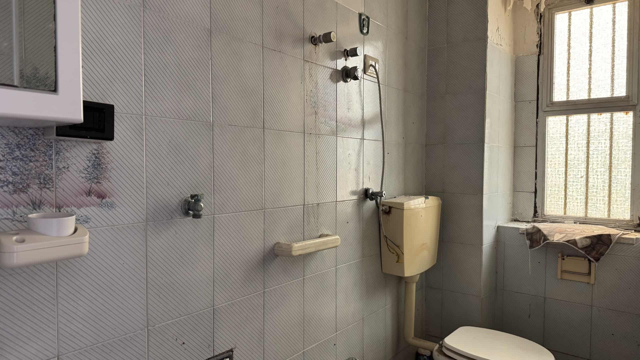 Doccia accessibile in un bagno con pareti rivestite di piastrelle grigie, maniglioni di sicurezza e finestra con grate, ideale per persone con esigenze di mobilità.