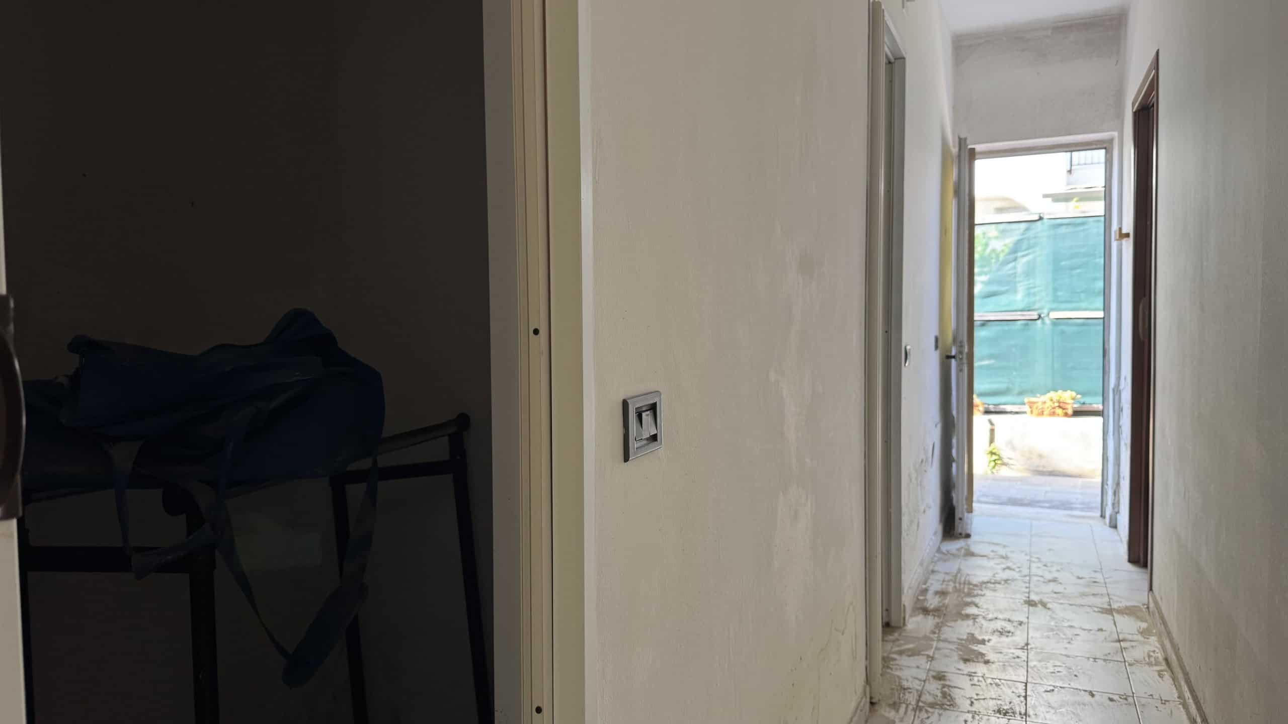 Corridoio interno di un appartamento in fase di ristrutturazione con pareti bianche e pavimento in piastrelle, vista verso l'esterno con porta aperta e luce naturale.