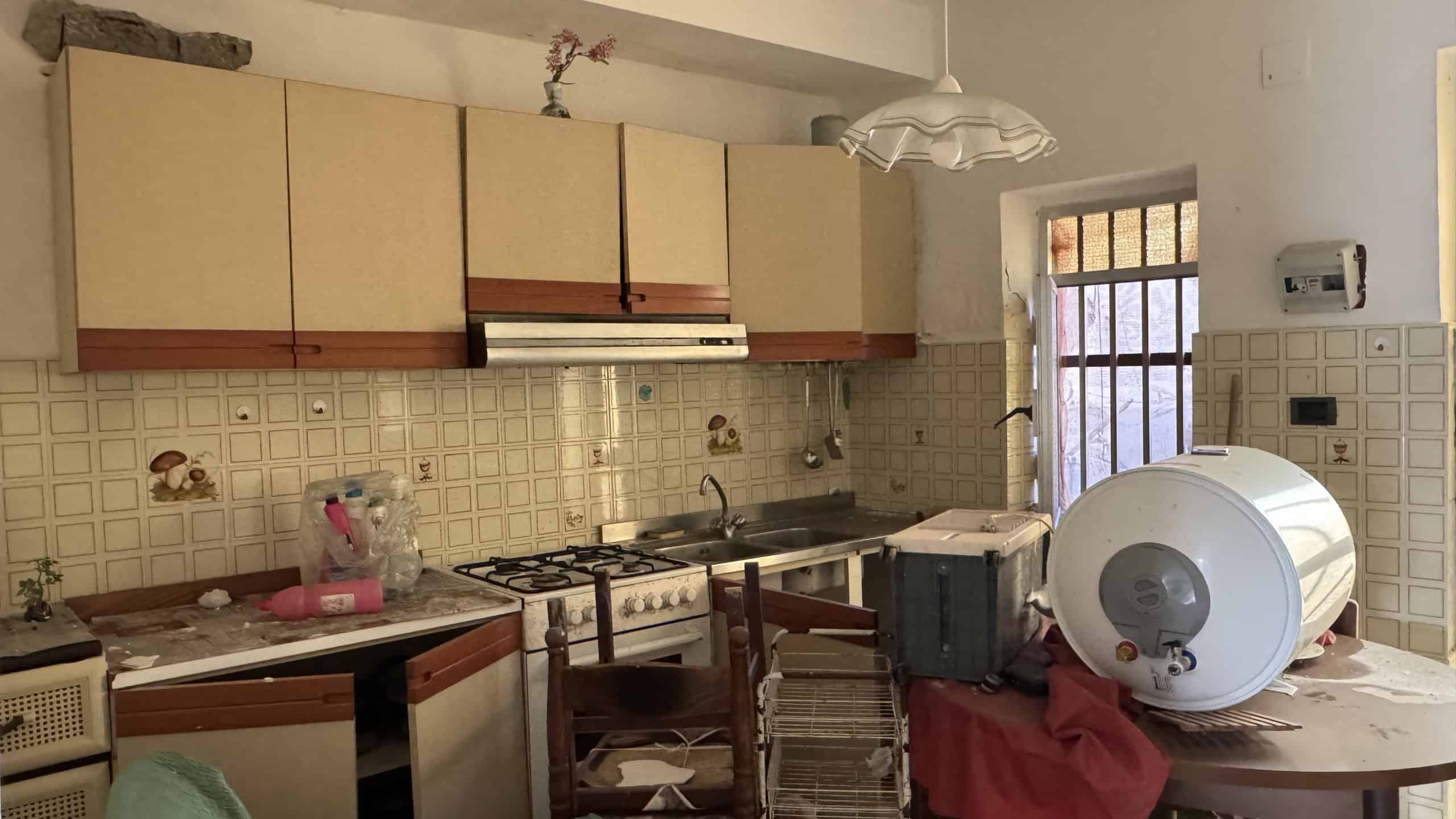 Spazio cucina abitabile con mobili classici, piano cottura a gas, lavandino e accessori vari, illuminata da luce naturale attraverso una finestra. Ambiente funzionale e accogliente, ideale per la prep.