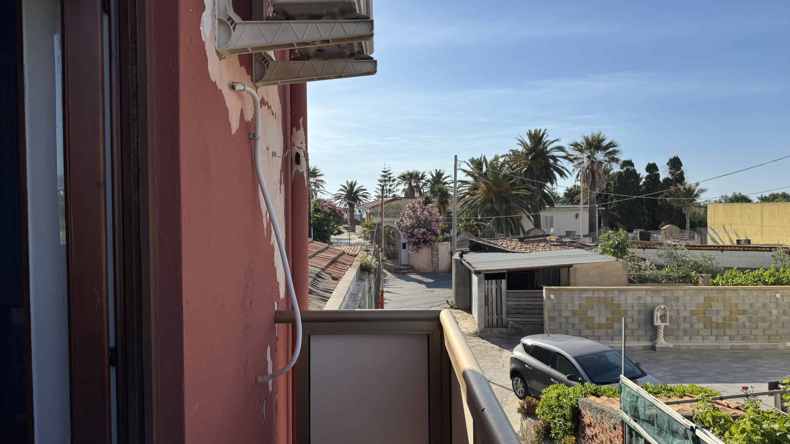Scorcio di un balcone con vista su strada urbana, palme e case in un quartiere residenziale soleggiato, atmosfera tranquilla e vivace.