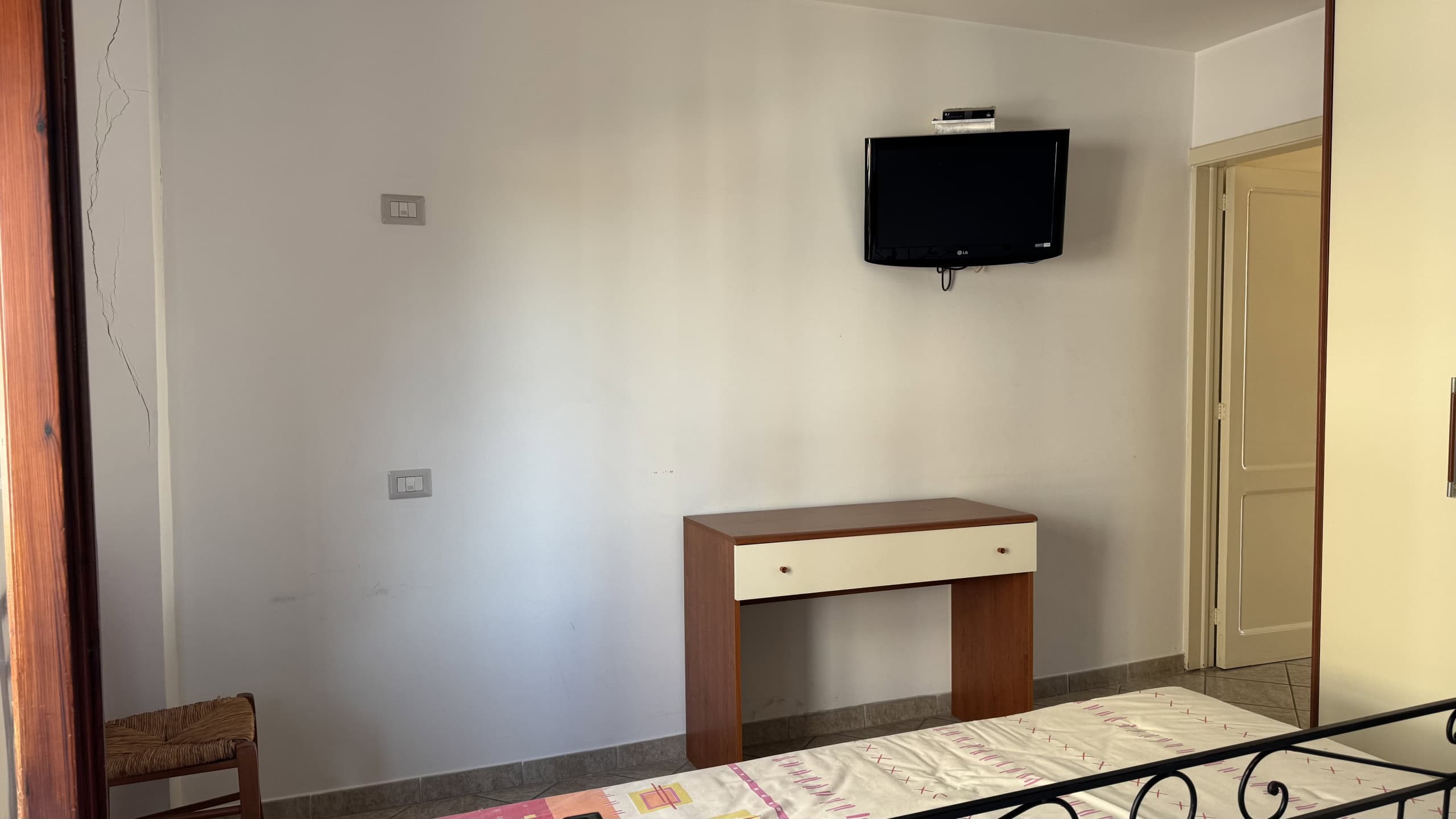 Interno di un soggiorno moderno con TV a parete e mobile in legno, pareti bianche e pavimento in piastrelle, ambiente luminoso e accogliente.