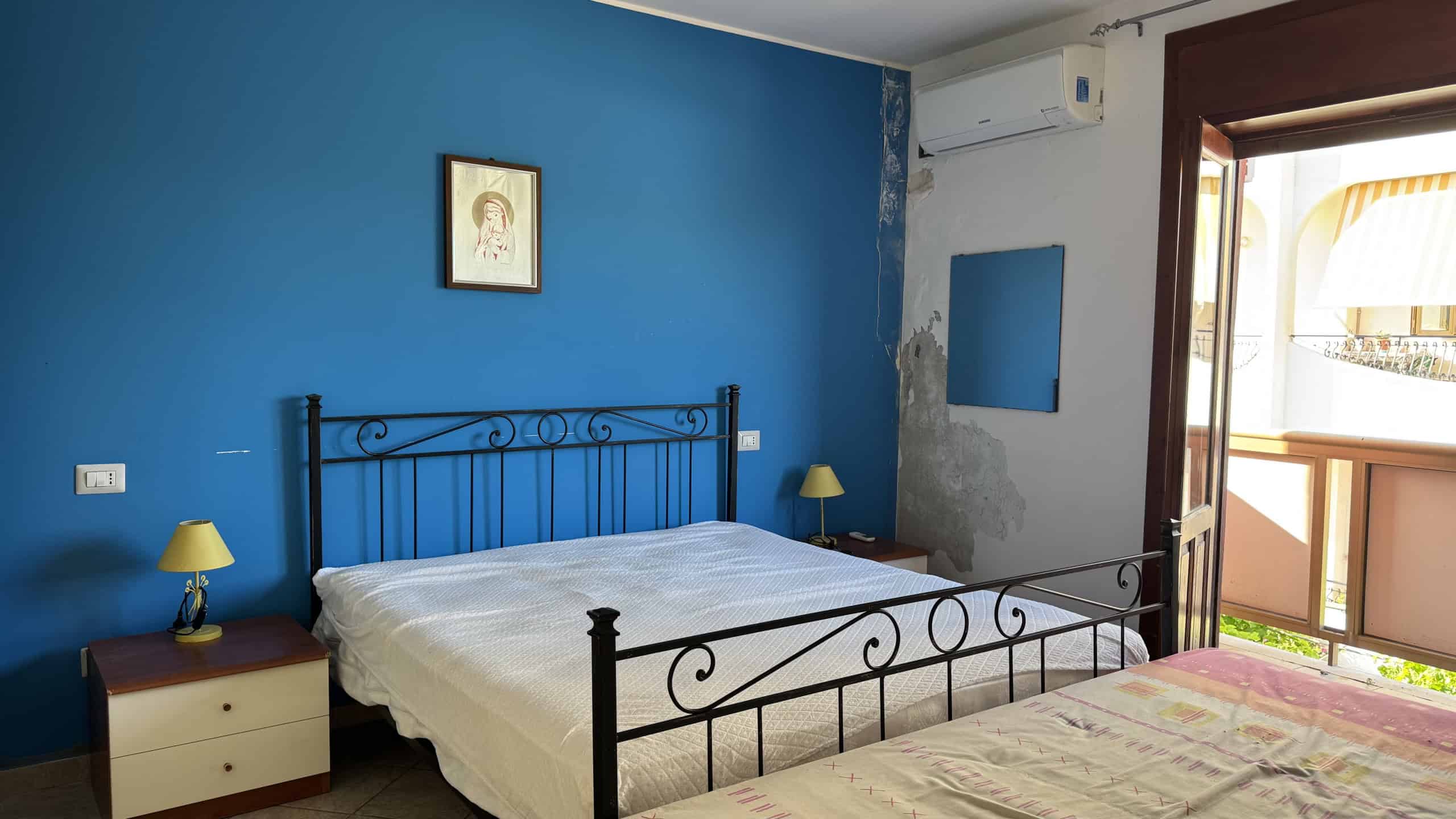 Camera da letto con pareti blu, letto in ferro battuto, balcone con vista, arredi semplici e funzionali.