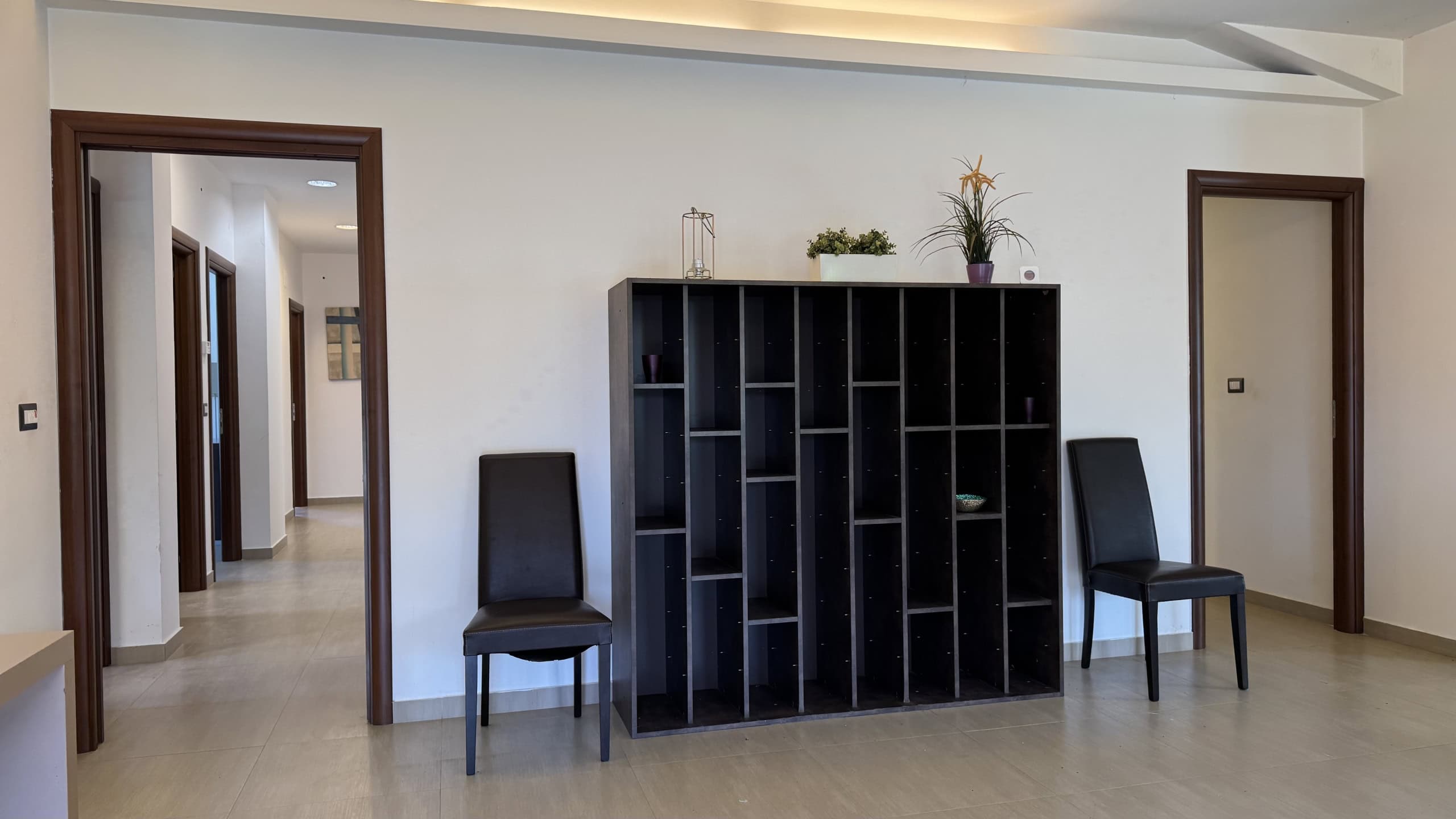 Interno di una villa unifamiliare con pareti bianche, pavimento in legno chiaro e due sedie nere ai lati di una libreria nera centrale.