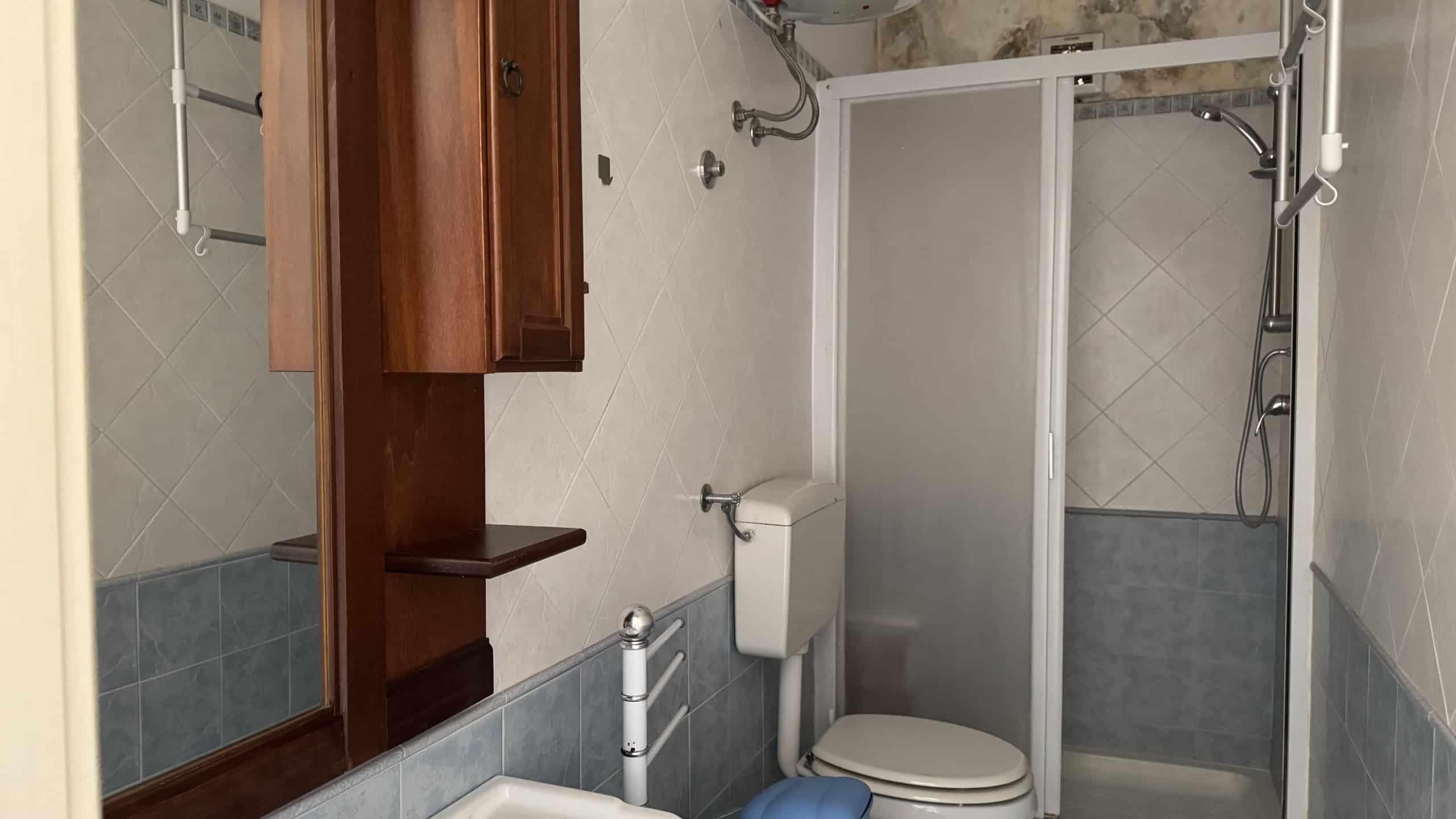 Bagno moderno ristrutturato con box doccia, sanitari e arredi in legno, pareti rivestite in piastrelle beige e azzurre, ideale per appartamenti in vendita o affitto a NextCasa.