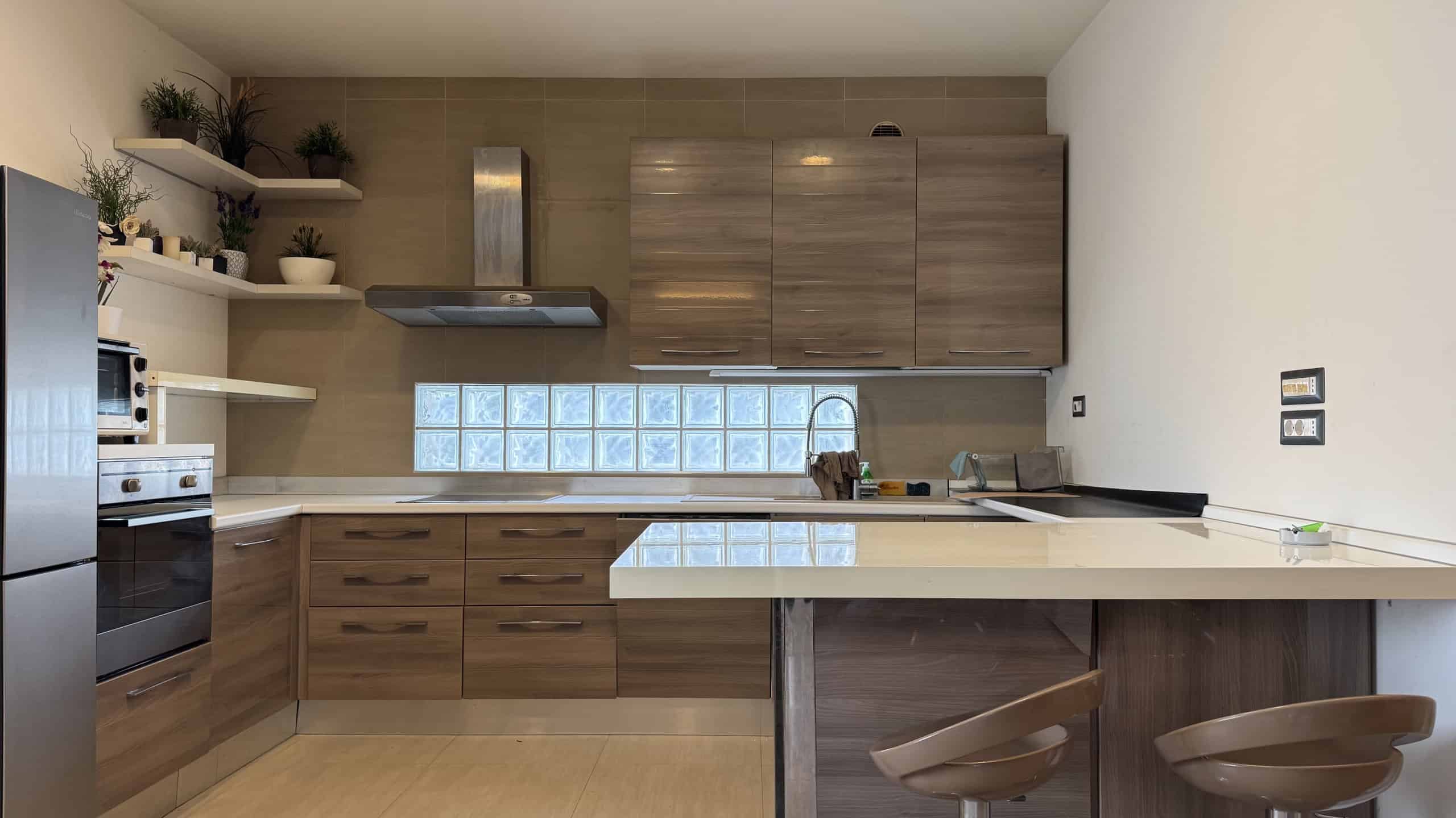 Cucina moderna con mobili in legno chiaro, piani in marmo e pareti bianche, con scaffali aperti decorativi e elettrodomestici integrati.