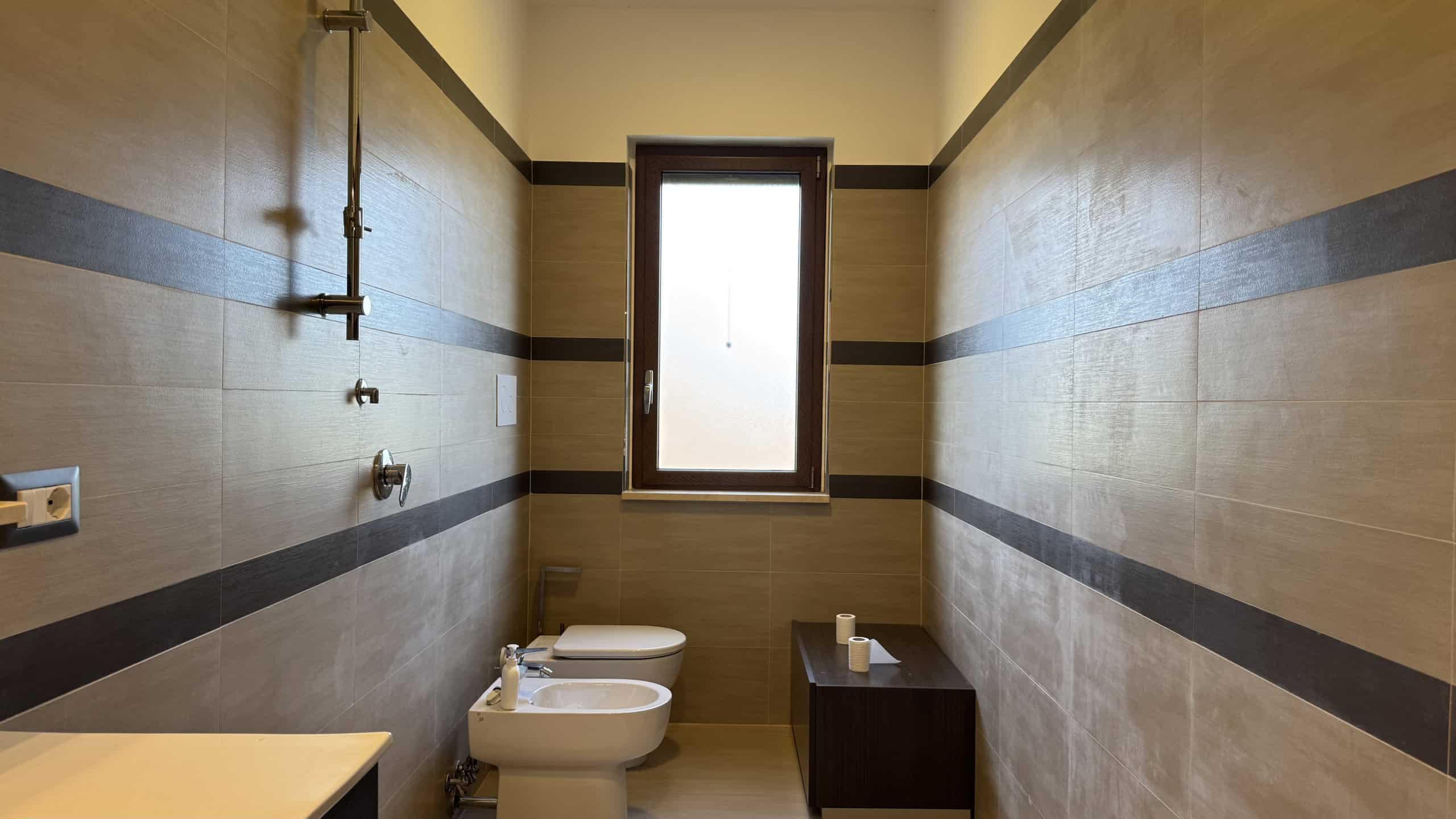 Interno di un bagno moderno con pareti beige e dettagli in nero, finestra centrale e sanitari bianchi, in una villa in Contrada Rosa Isolera 7, Corriolo, Me.