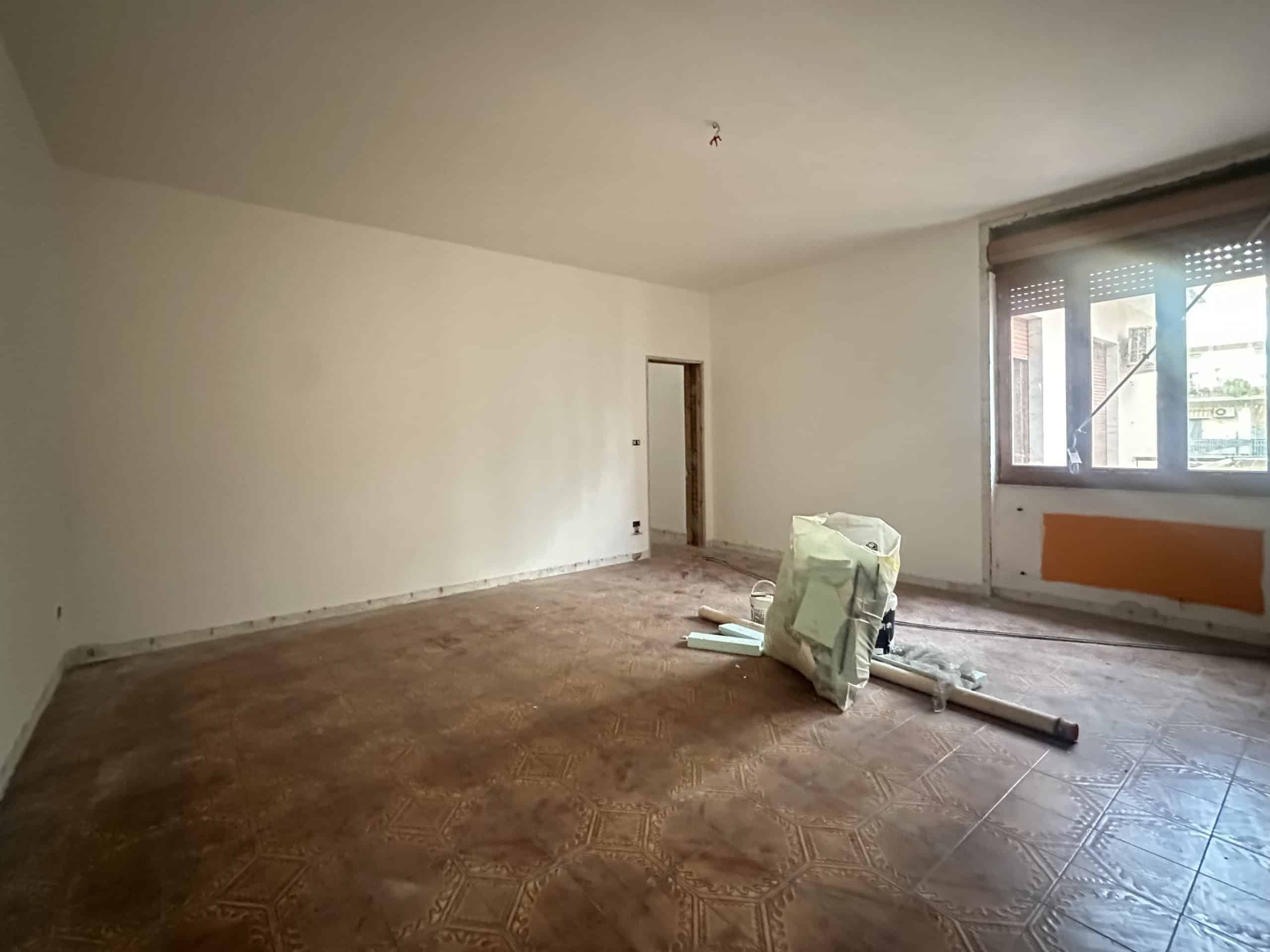 Spazio interno di un appartamento in fase di ristrutturazione con pareti bianche e pavimento in piastrelle, finestra grande e porta aperta.