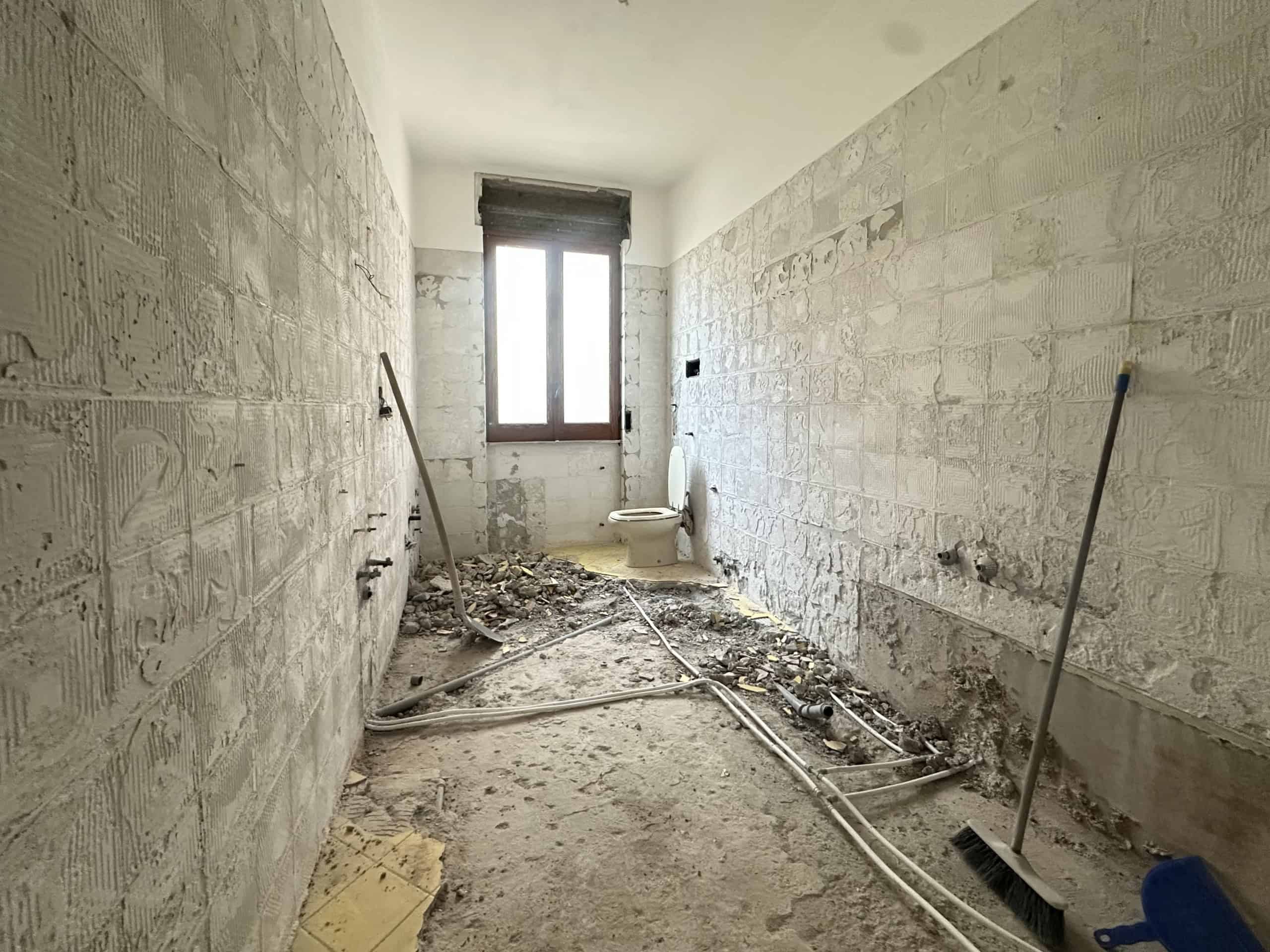 Interno di una stanza in ristrutturazione con pareti in mattoni e tubature visibili, finestra grande sulla parete opposta.
