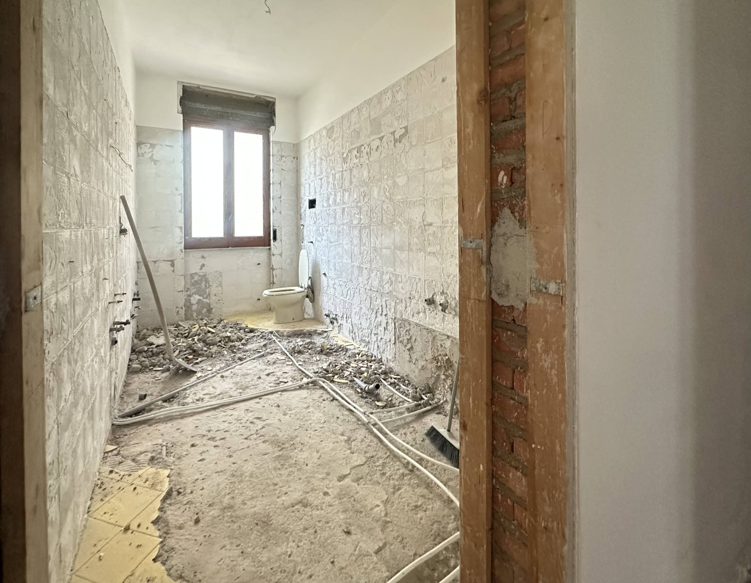 Interno di un bagno in fase di ristrutturazione con pareti in muratura e tubature visibili, luce naturale dalla finestra.
