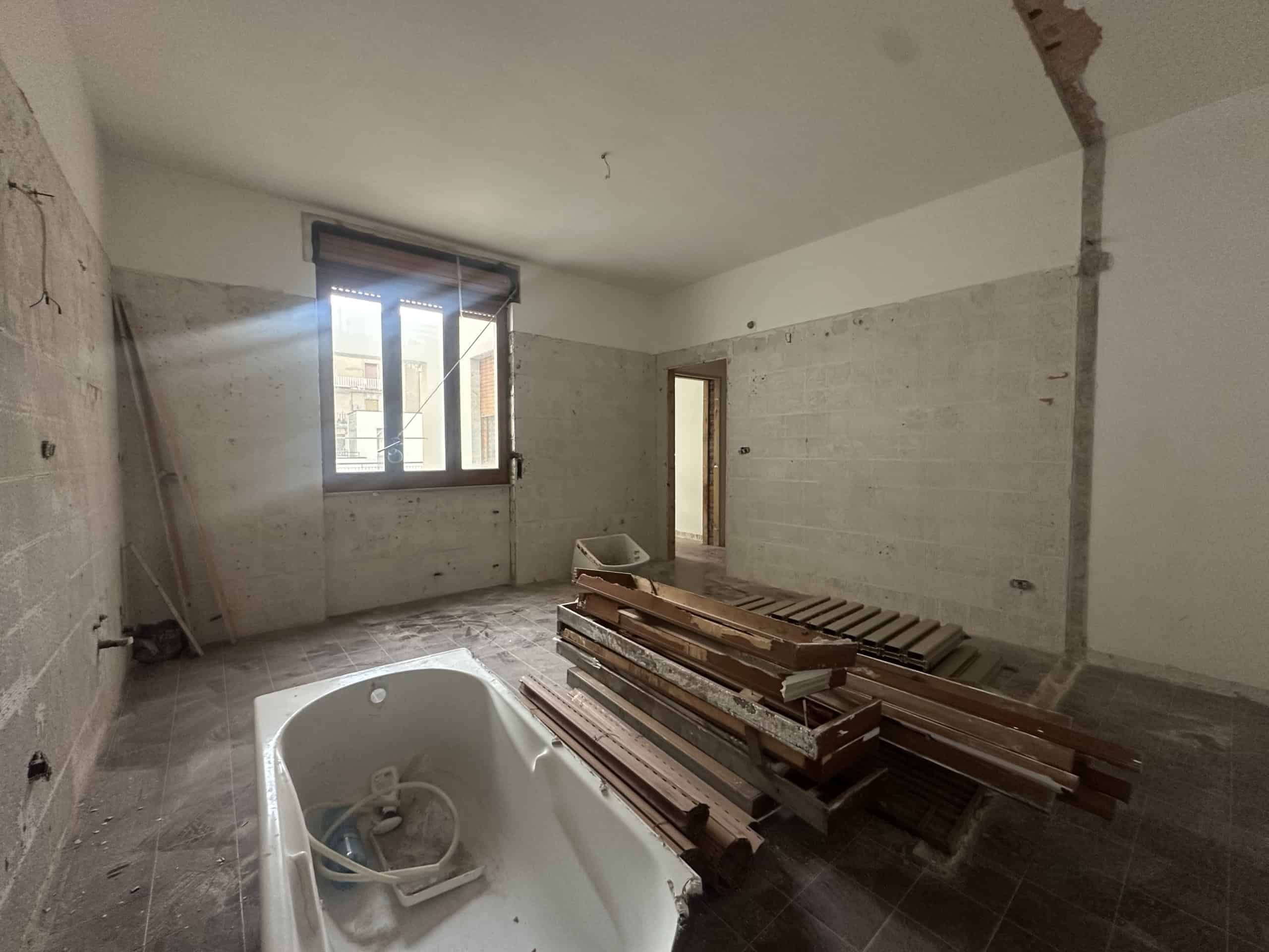 Interno di una stanza in ristrutturazione con finestra, pareti in cemento e materiali da costruzione sul pavimento.