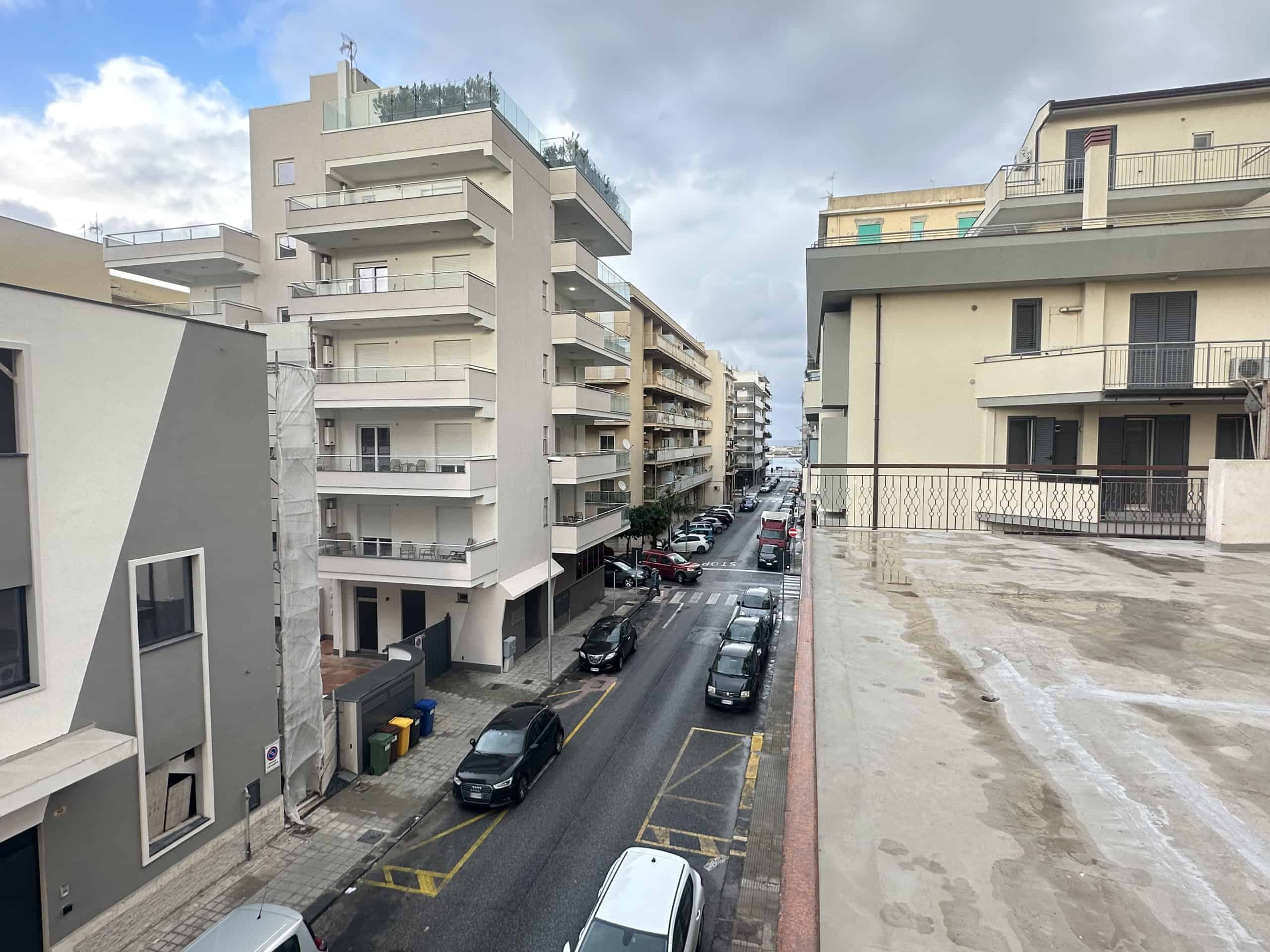 Immagine di un quartiere residenziale con edifici moderni e strada trafficata, con auto parcheggiate e cielo nuvoloso.