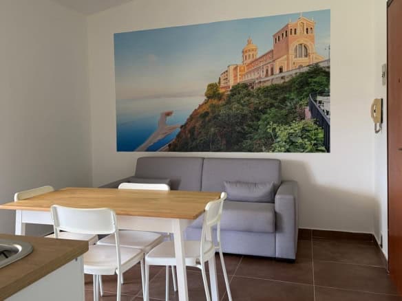 Interno di un appartamento con soggiorno, divano grigio, tavolo in legno e sedie bianche, decorato con grande foto di un paesaggio costiero sulla parete.