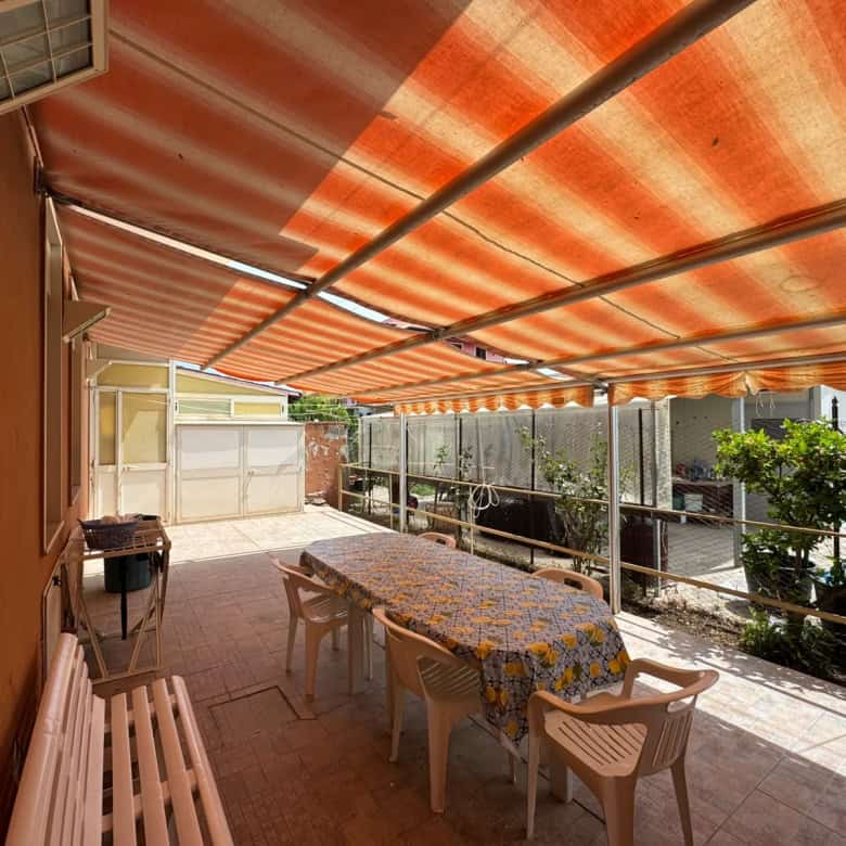 Terrazza con tavolo e sedie sotto tenda arancione, vista su giardino e case circostanti.