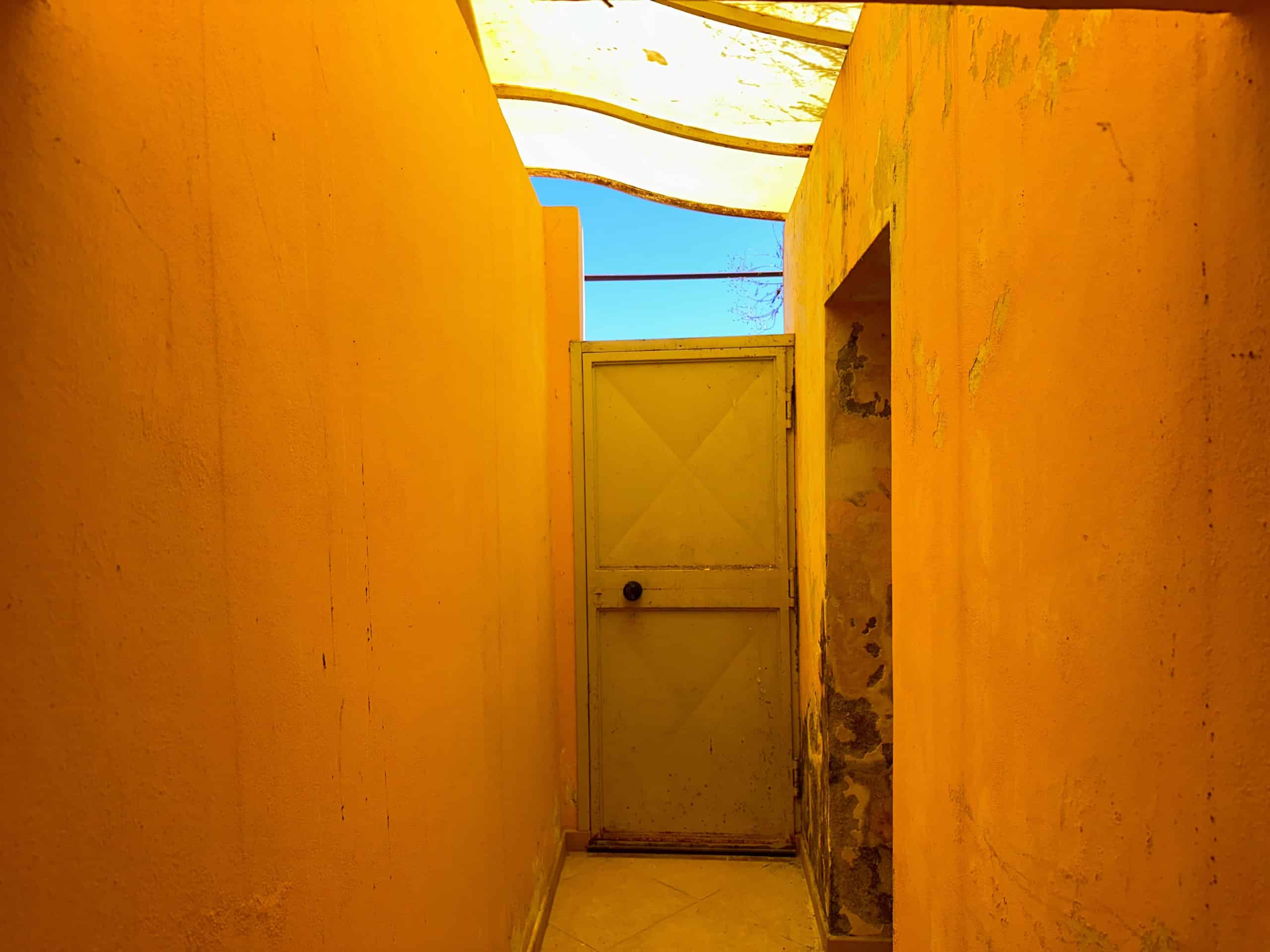 Ingresso di un cortile interno di una casa con pareti gialle e porta bianca, sotto un cielo azzurro.