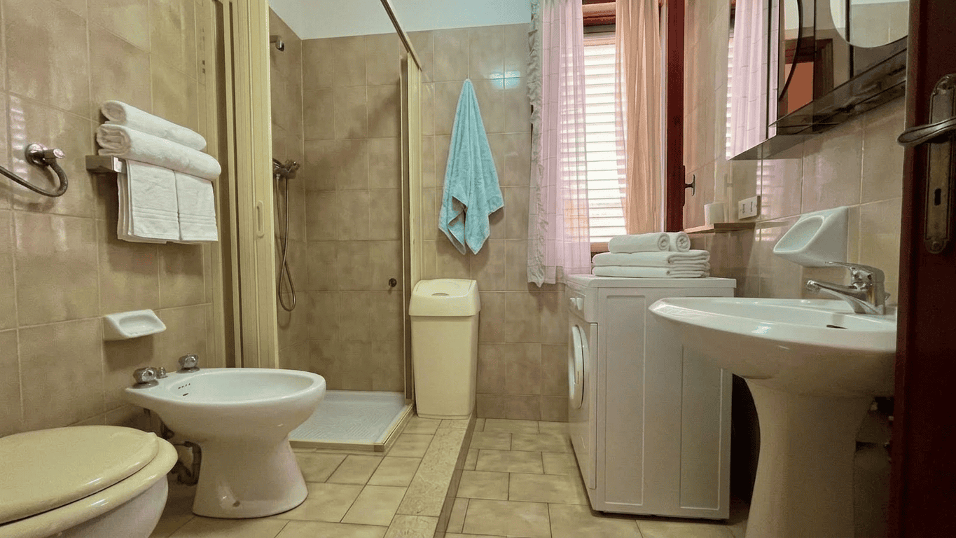 Bagno spazioso con lavabo, bidet, WC, lavatrice e doccia, arredato con tende rosa e asciugamani bianchi, in un appartamento in vendita a Oliveri, Me.