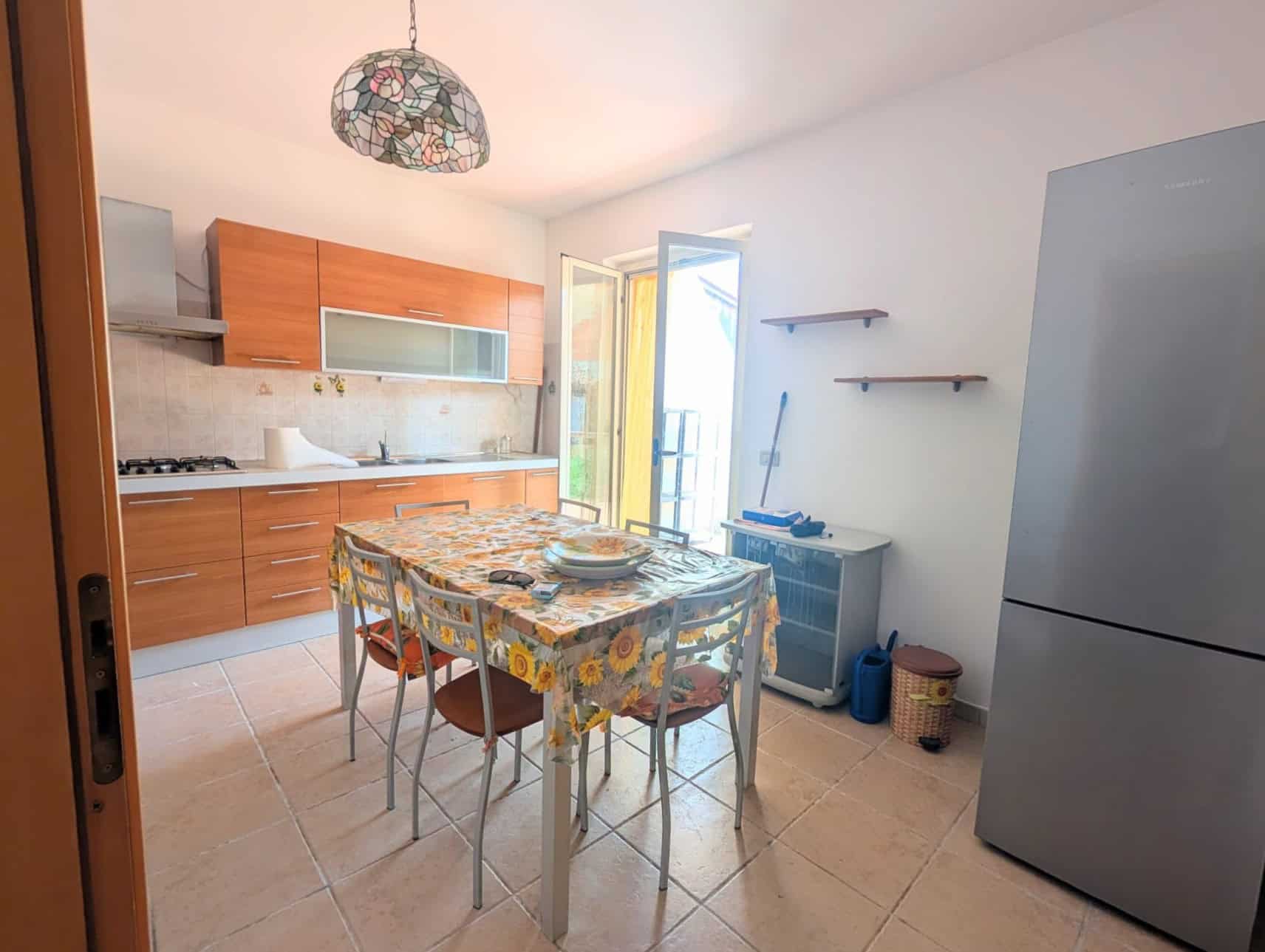 Cucina luminosa con mobili in legno, tavolo con tovaglia floreale e frigorifero in una villetta a schiera.