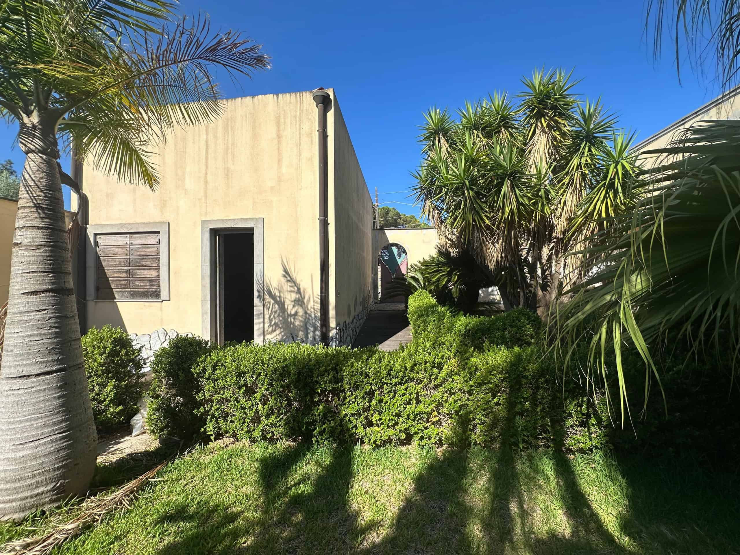 Giardino con piante tropicali e siepi, casa in stile moderno in background, cielo sereno.